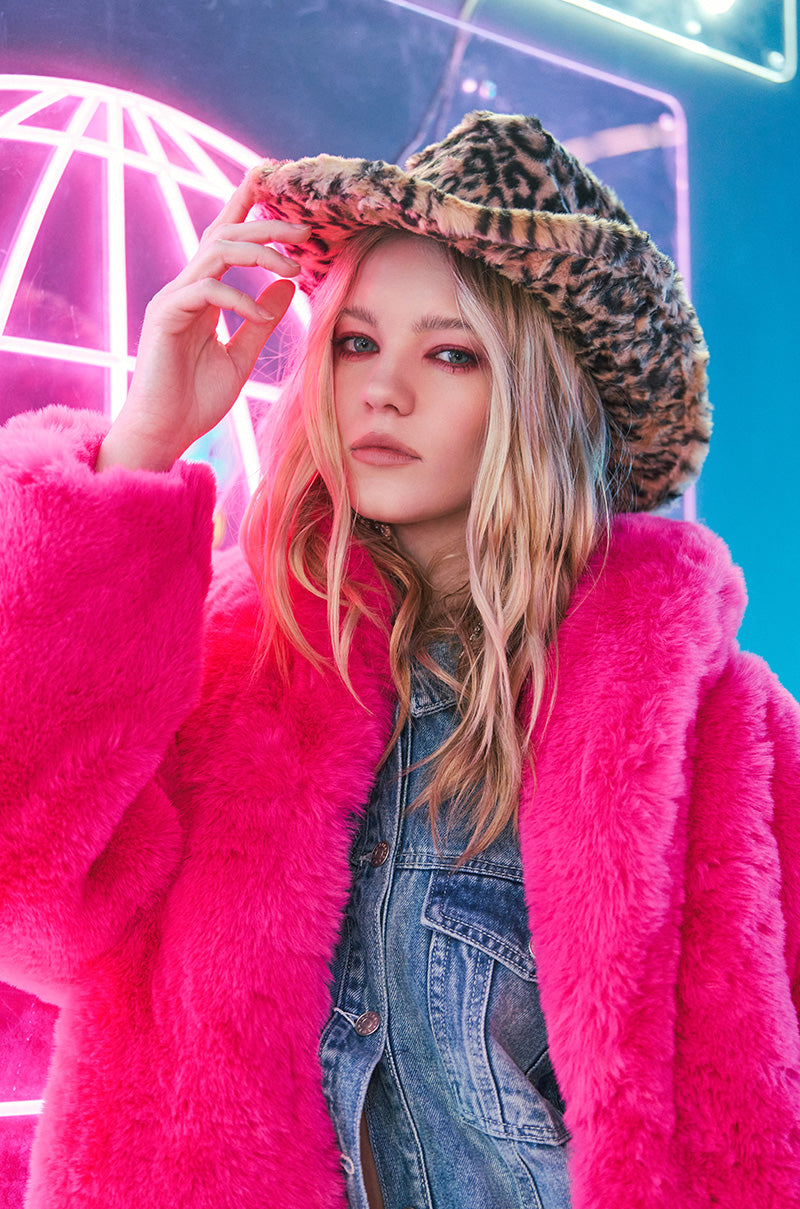 Wearing a vibrant pink faux fur coat and denim outfit, a person adjusts the CHECK ME OUT FAUX FUR COWBOY HAT with its striking leopard-print design. In the background, neon pink lights shaped like a globe enhance the colorful, modern aesthetic with hints of Western flair.