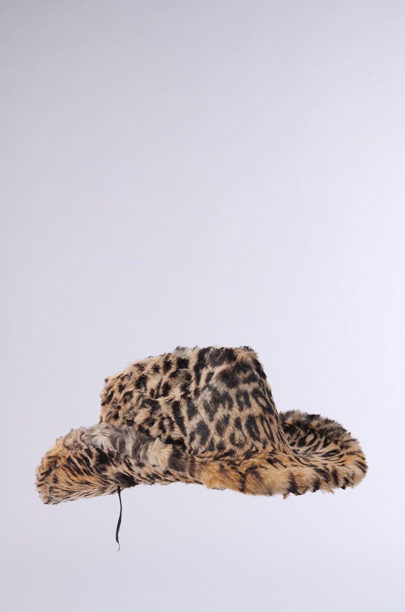 The CHECK ME OUT FAUX FUR COWBOY HAT, featuring a leopard print pattern with a slightly floppy brim and a black string hanging from the side, is showcased against a plain white background, making it an ideal Western accessory.