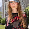 A young woman with long, wavy hair wears the IT GURL PU BRIM HAT in gray and a black mesh top with red sequined "AUTHENTIC 25" text, outdoors in a city park with skyscrapers behind her.