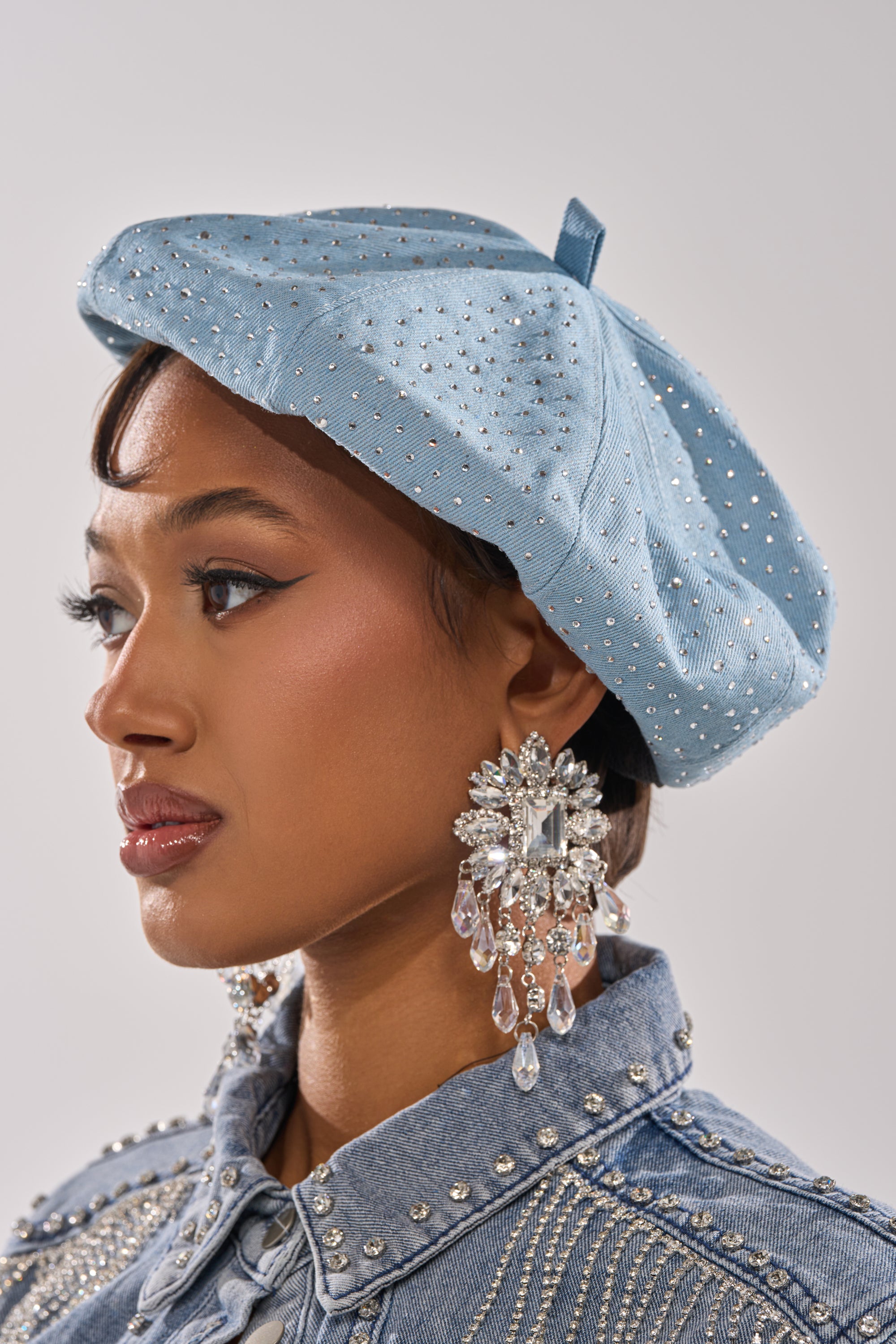 A woman in the SEE ME COMING BLING BERET, a light blue rhinestone hat, pairs it with a matching denim jacket with rhinestone accents and large crystal earrings, looking to the left against a plain background.