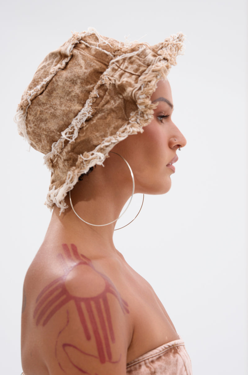 A person in profile wears the WITH IT DISTRESSED BUCKET HAT in beige, paired with large hoop earrings, a nose ring, and a bold red abstract shoulder tattoo. The background is plain white.