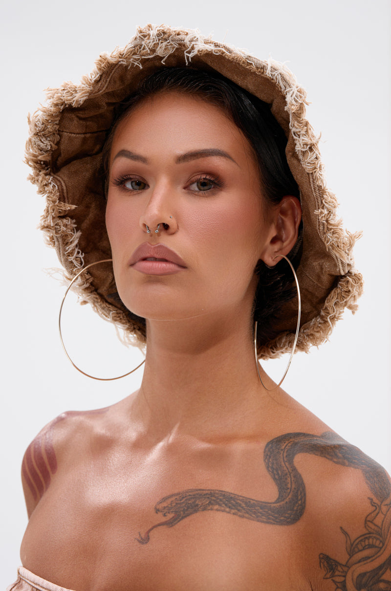 Wearing the WITH IT DISTRESSED BUCKET HAT, a woman with large hoop earrings, a nose ring, and a snake tattoo on her shoulder looks confidently at the camera against a plain background.