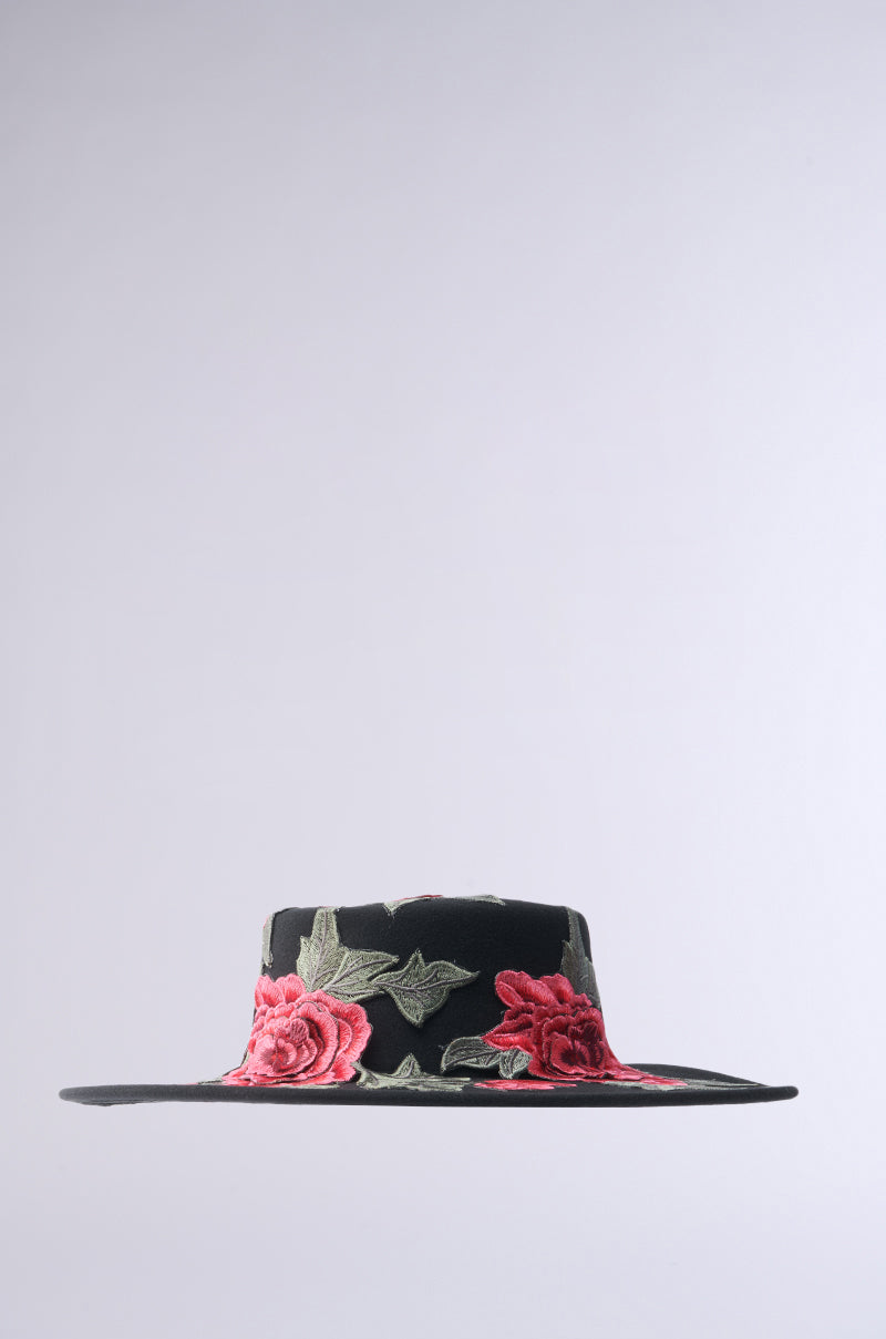 The ALIYA FLORAL BRIM HAT is a black wide-brimmed hat with a flat top, featuring embroidered red roses and green leaves. Its plain light gray background highlights the intricate floral details. 6