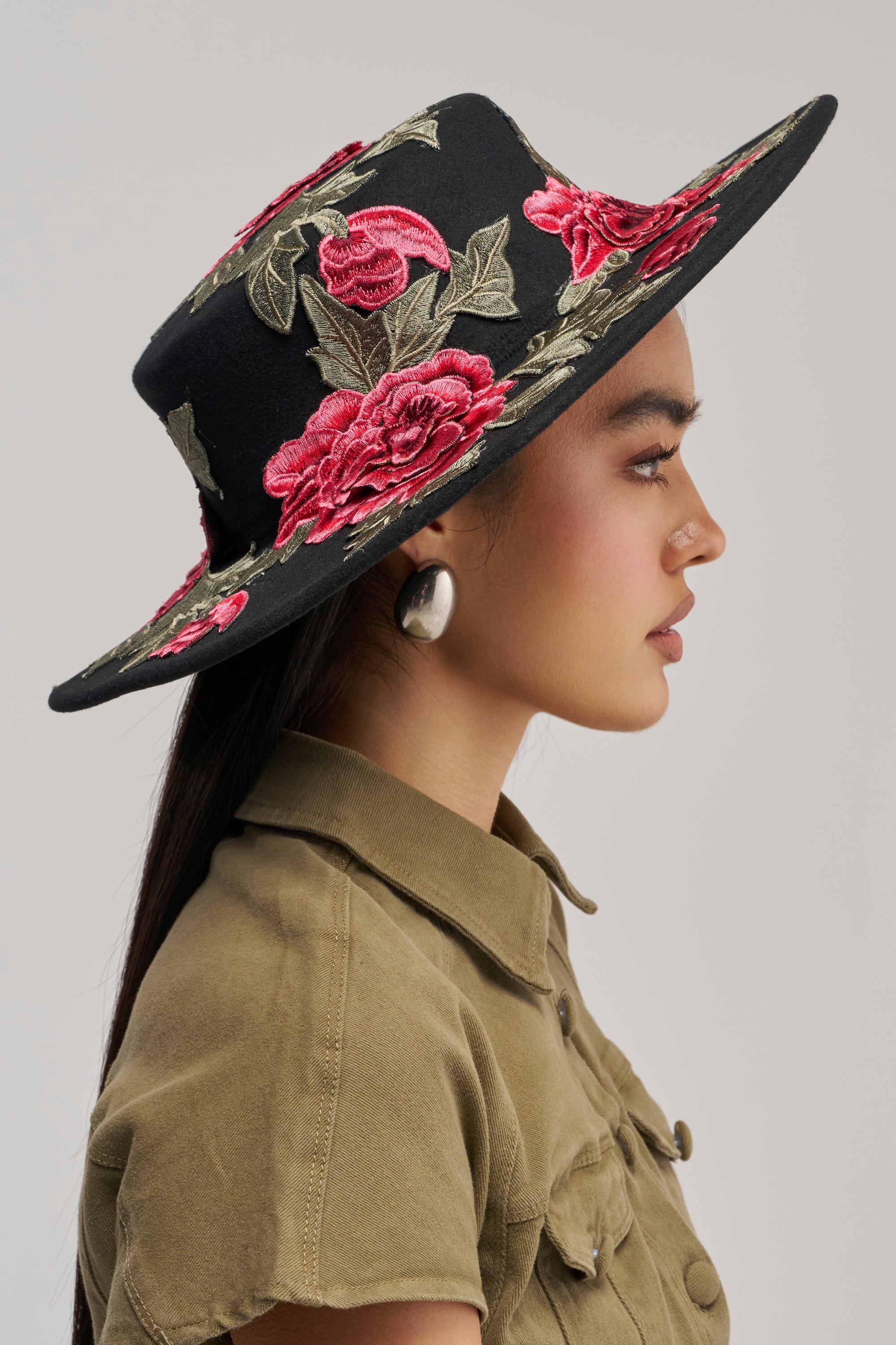 A woman in profile wears the ALIYA FLORAL BRIM HAT, a striking black hat embroidered with roses and featuring a wide brim. She pairs it with large round earrings and a khaki collared shirt—perfect inspiration for festival accessorizing. The background is plain and light-colored.
