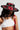 A woman in the ALIYA FLORAL BRIM HAT, with floral appliqués, gazes down. Her long dark hair flows over a white crop top, adorned with a pearl necklace and a red leather strap with metal studs. The scene unfolds against a plain gray backdrop, exuding AKIRA Label charm.