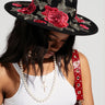 A woman in the ALIYA FLORAL BRIM HAT, with floral appliqués, gazes down. Her long dark hair flows over a white crop top, adorned with a pearl necklace and a red leather strap with metal studs. The scene unfolds against a plain gray backdrop, exuding AKIRA Label charm.