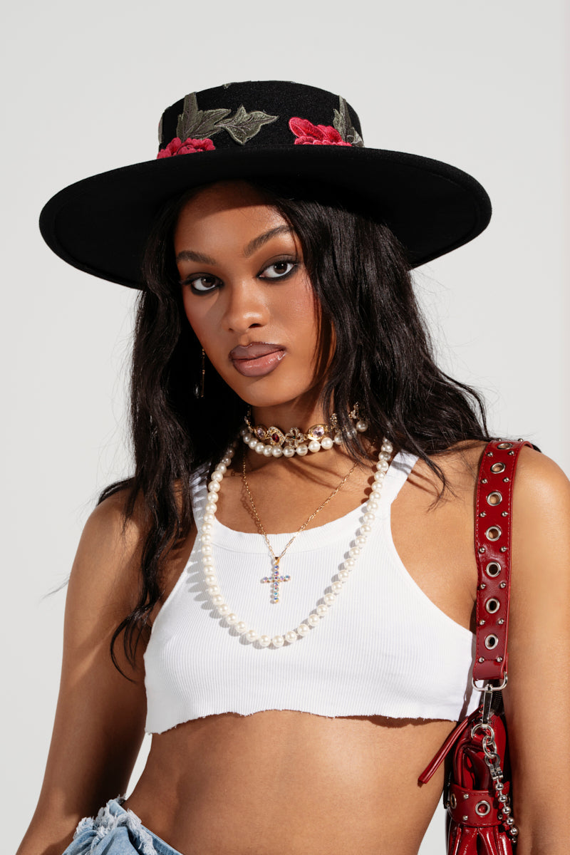 A woman wears an ALIYA Floral Brim Hat with embroidered floral appliqués, a white cropped tank top, layered pearl necklaces, and a red AKIRA Label purse with metal accents. Her long dark hair flows as she exudes confidence. 4