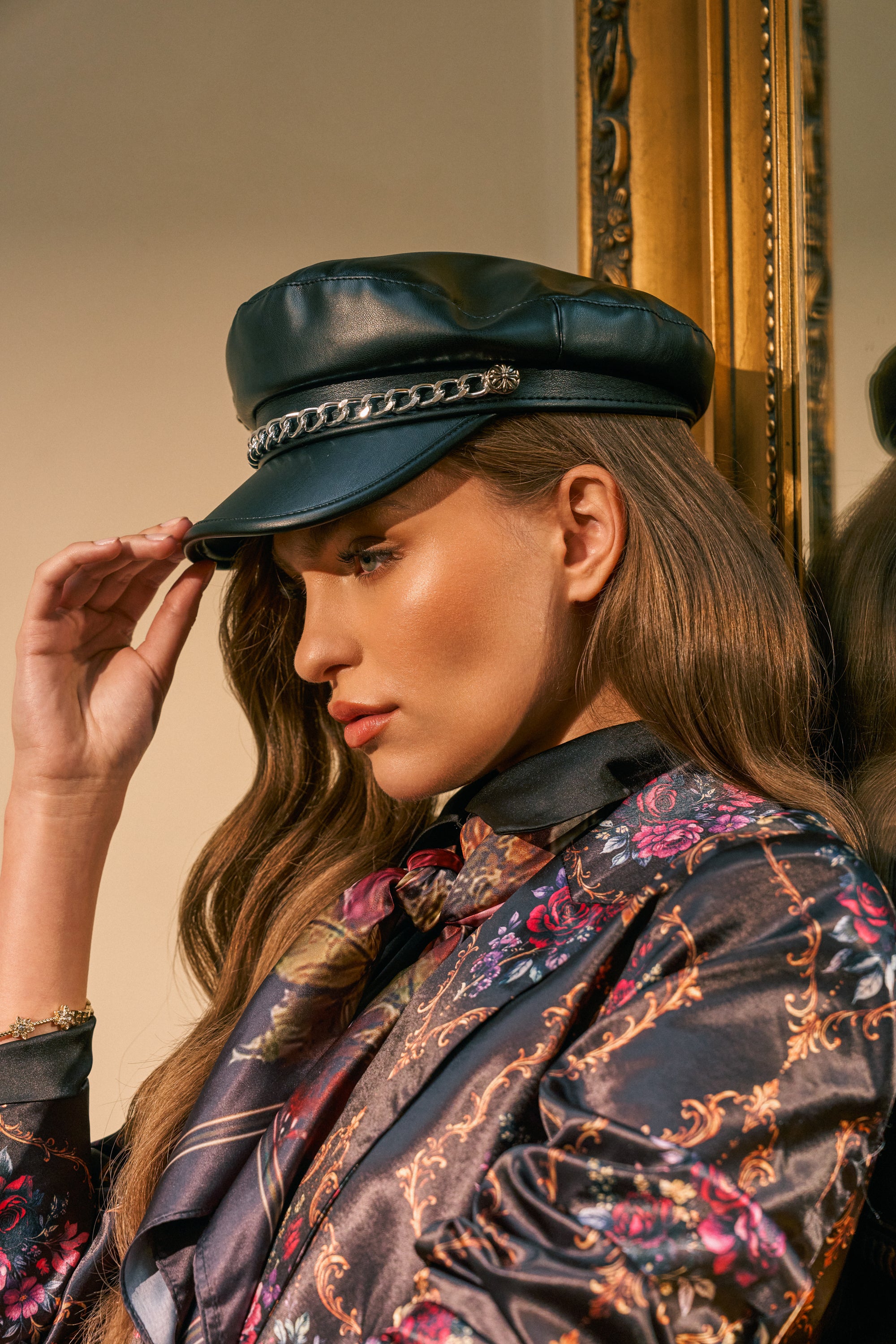 A person with long wavy hair wears the CROSS ME BRIM HAT, a black faux leather cabbie hat accented with a chain detail, paired with a floral patterned jacket. Standing next to a gold-framed mirror, they appear thoughtful.