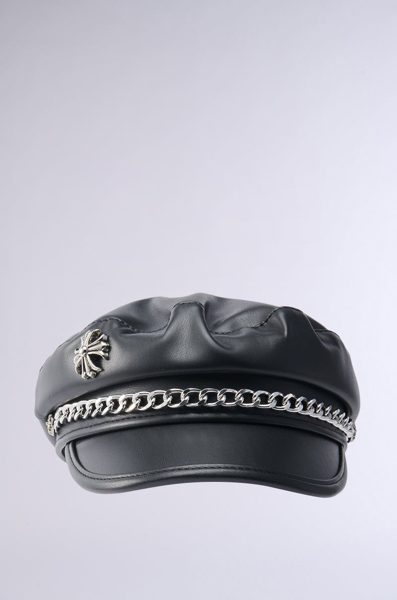 Introducing the CROSS ME BRIM HAT, a standout accessory in sleek faux black leather, featuring a unique silver metallic chain along the brim and a polished silver emblem on its side, beautifully contrasted against a plain light gray background.