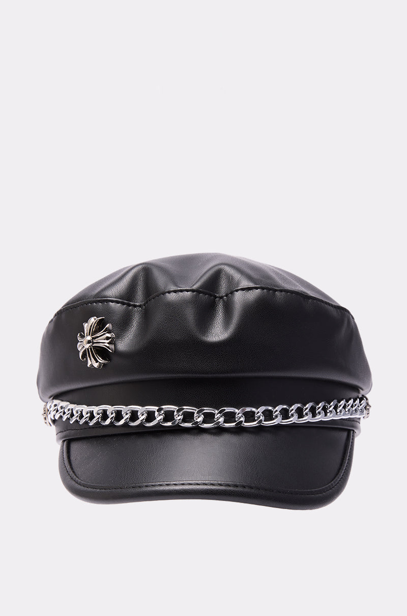 The CROSS ME BRIM HAT is a black faux leather hat featuring a structured, glossy brim, a shiny silver chain detail across the front, and a metal spider-shaped pin on the left side. 4