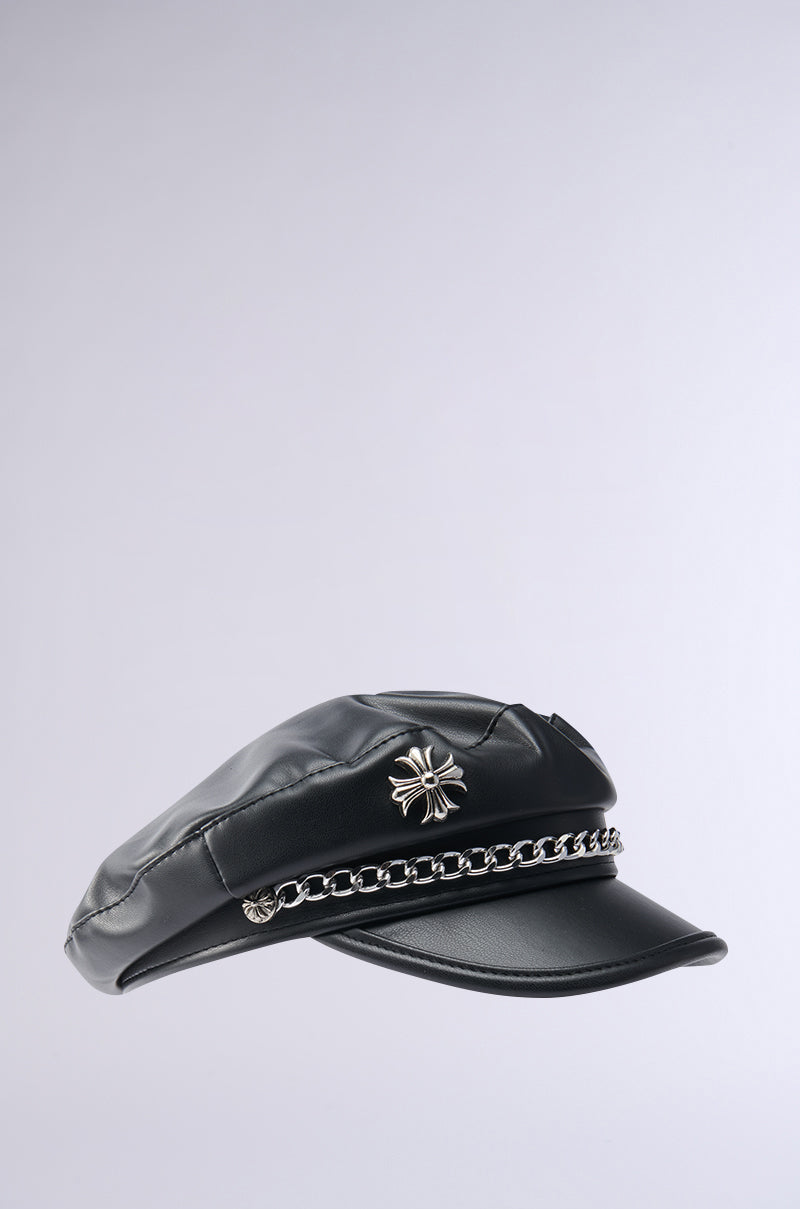 The CROSS ME BRIM HAT boasts sleek black faux leather, a silver metallic chain link above the brim, and a stylish silver emblem on the front, all contrasted against a light gray background.