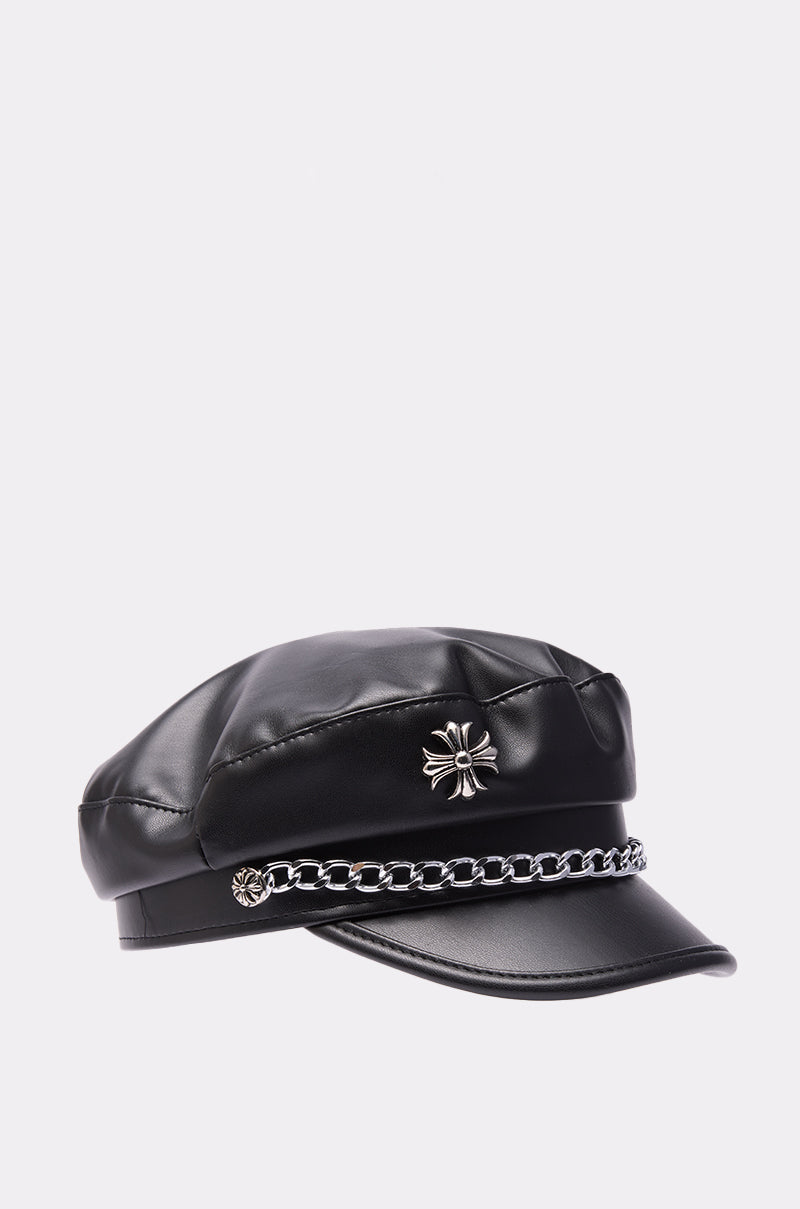The CROSS ME BRIM HAT is a black faux leather newsboy cap with a shiny silver chain link across the front and two silver flower accents, shown against a plain light background. 5