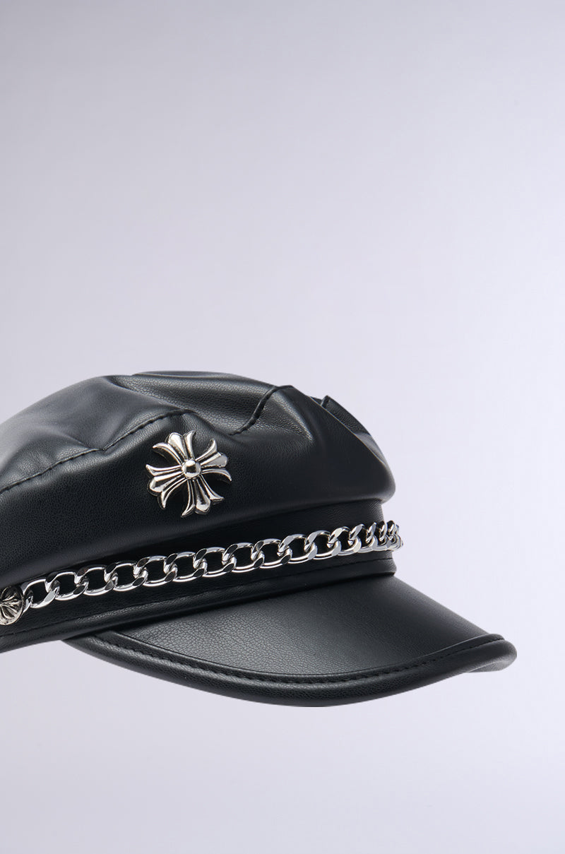 The CROSS ME BRIM HAT in black faux leather showcases a bold silver emblem with a silver metallic chain link on the front, set against a light gray backdrop.