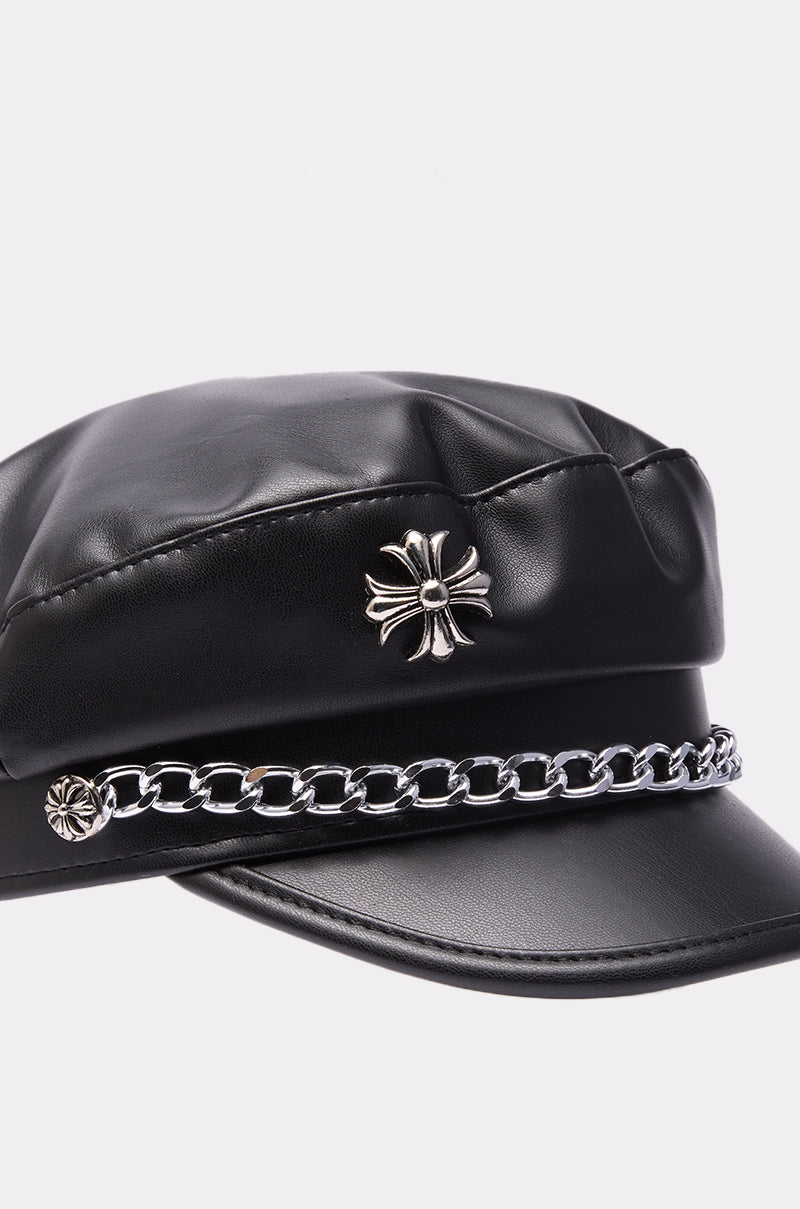 The CROSS ME BRIM HAT is a black faux leather hat featuring a shiny metallic chain link across the front and a silver floral emblem above the brim, displayed against a plain light background.