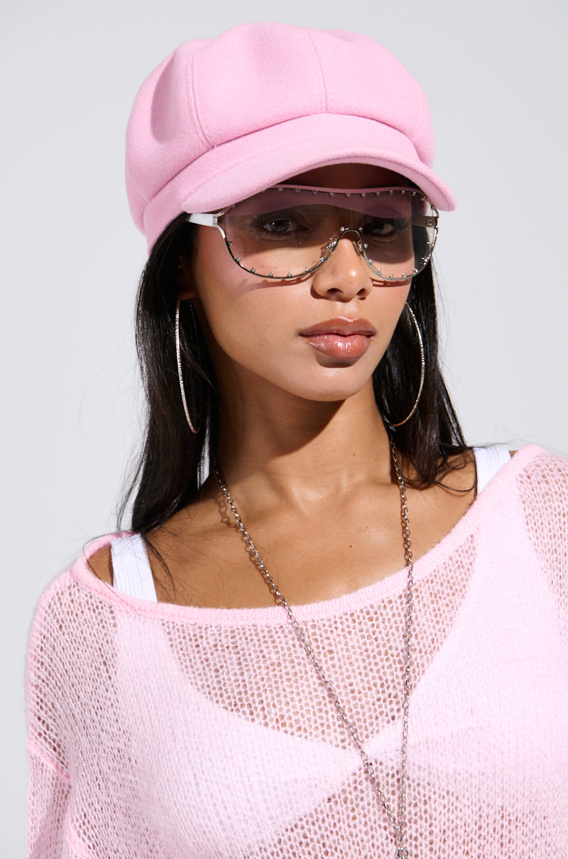 A person confidently poses in a PRETTY IN PINK BRIM HAT and sheer pink top, accessorized with large sunglasses, hoop earrings, and a chain necklace. The style is casually trendy against a neutral background.
