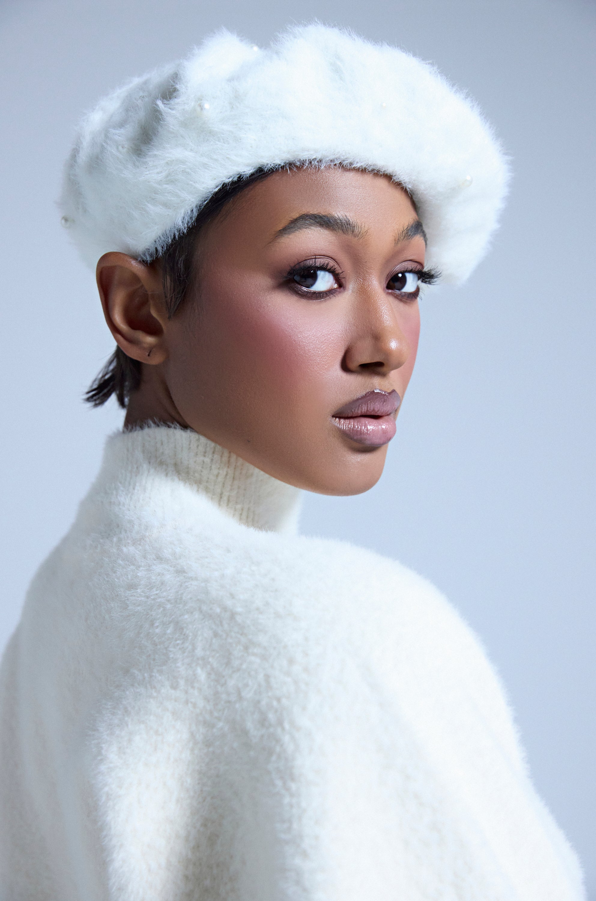 A woman wearing the PEARLY WHITES FLUFFY BERET, featuring pearl embellishments, and a matching knit sweater looks over her shoulder with soft makeup and a calm expression against a light background. 1