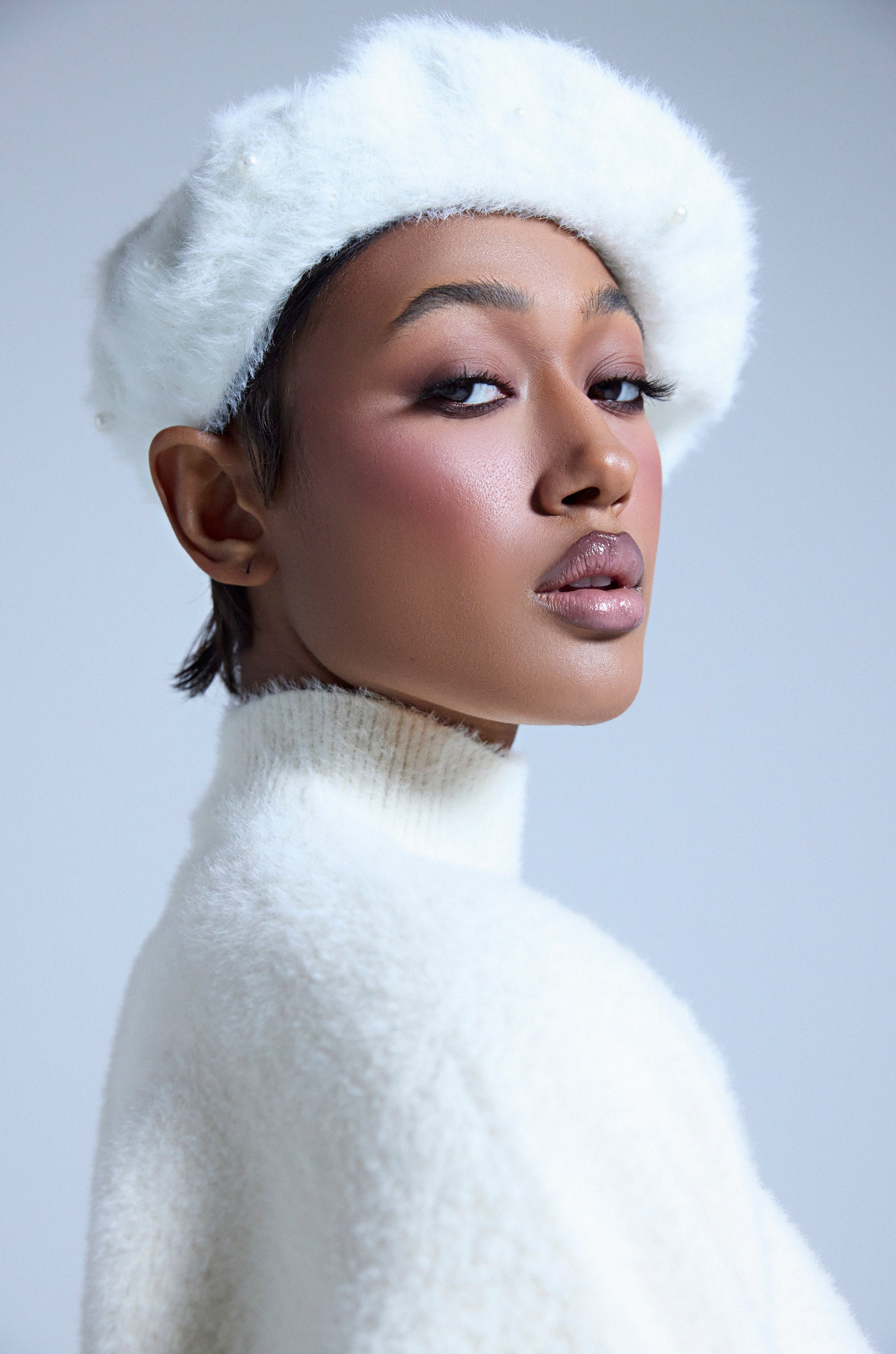 A woman wearing the PEARLY WHITES FLUFFY BERET and a white high-neck sweater looks confidently over her shoulder. The soft, light gray background highlights the beret’s clean and elegant style. 2