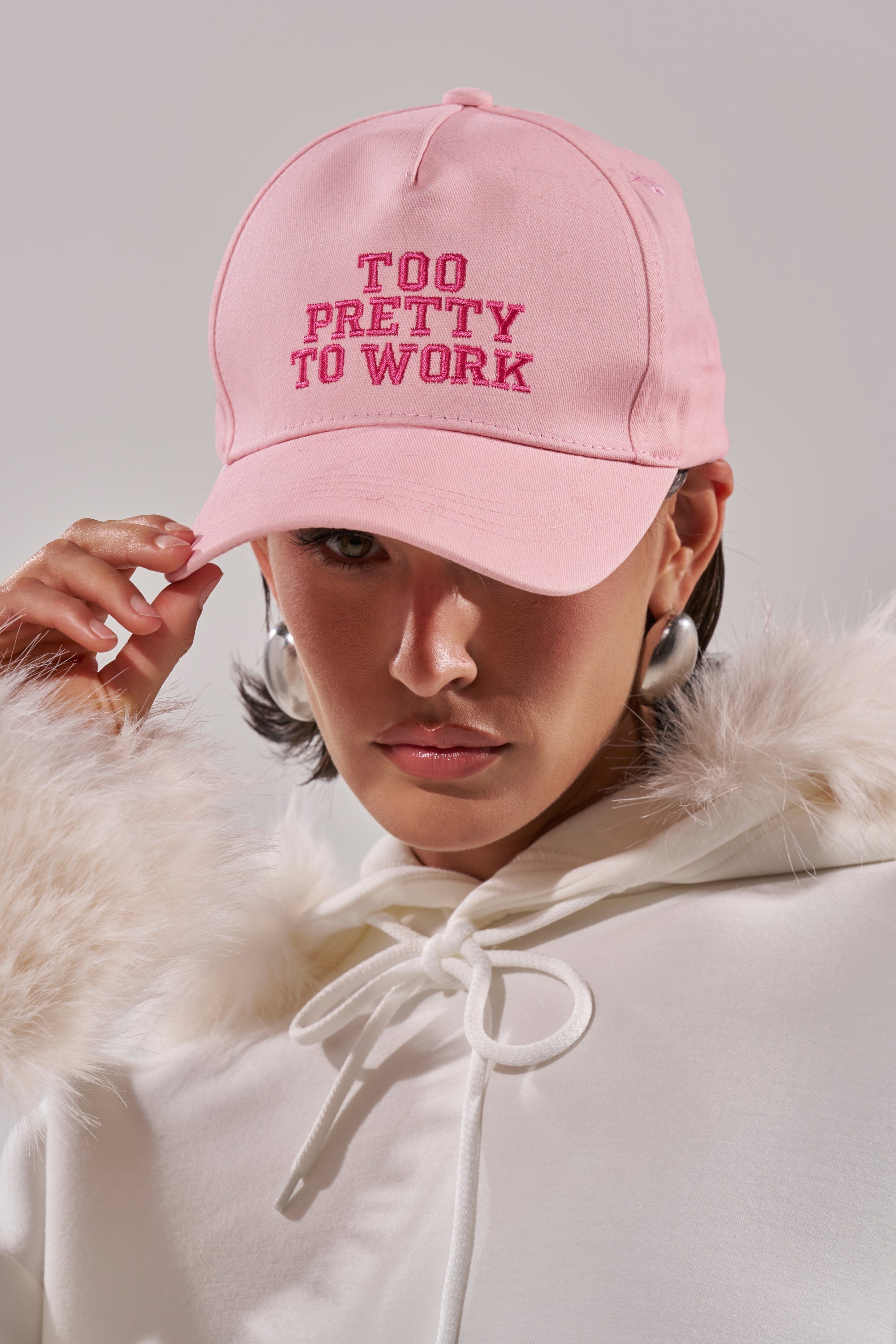 A person wears the I SAID WHAT I SAID HAT in pastel pink, featuring embroidery, paired with a white fur-trimmed hoodie and bold hoop earrings, posing against a neutral background. 0