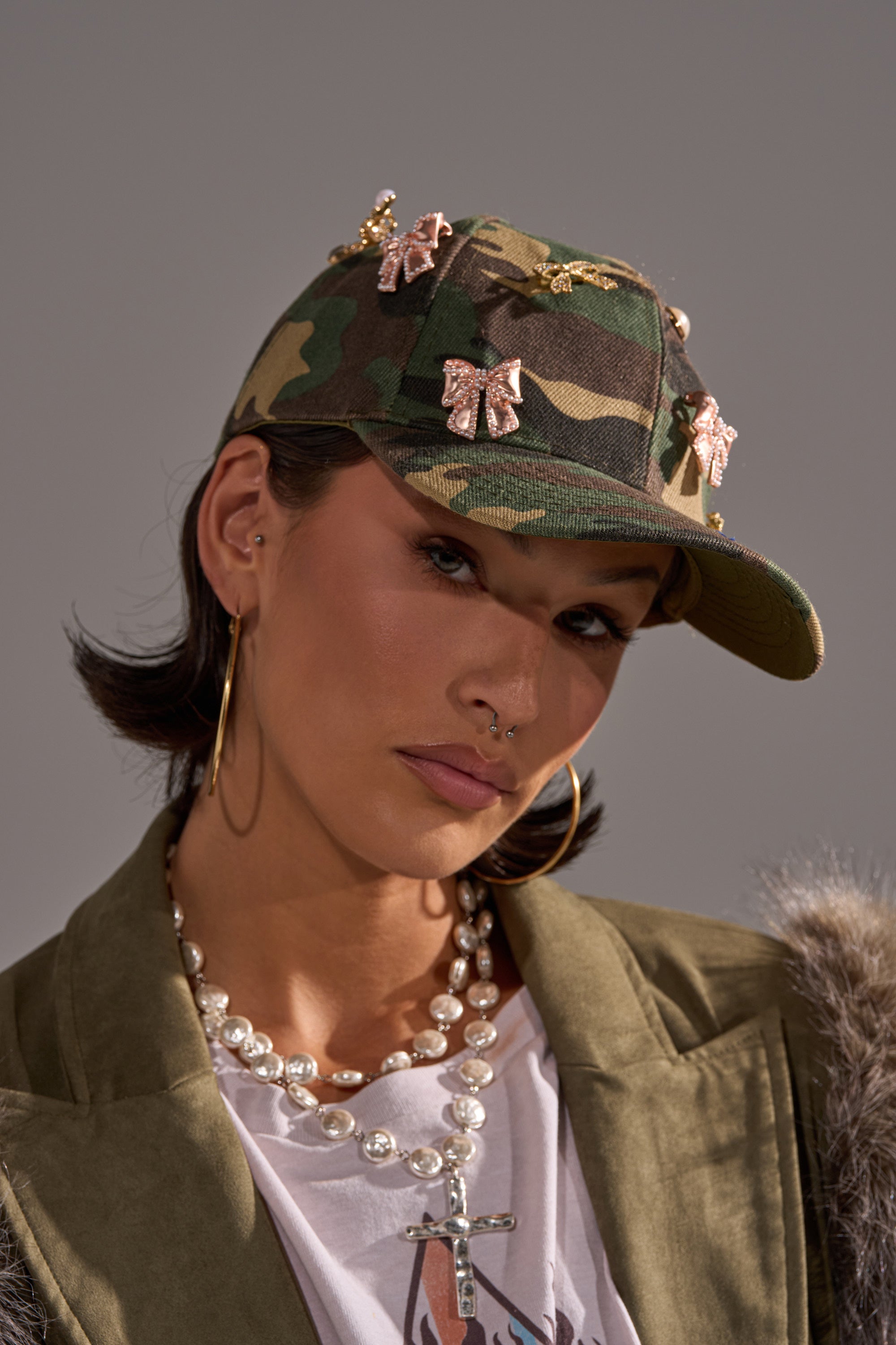 A woman wears the TANYA CHARM HAT, featuring a camouflage print and butterfly pins, paired with gold hoop earrings, a pearl cross necklace, a white tee, and an olive faux fur collar jacket. 0
