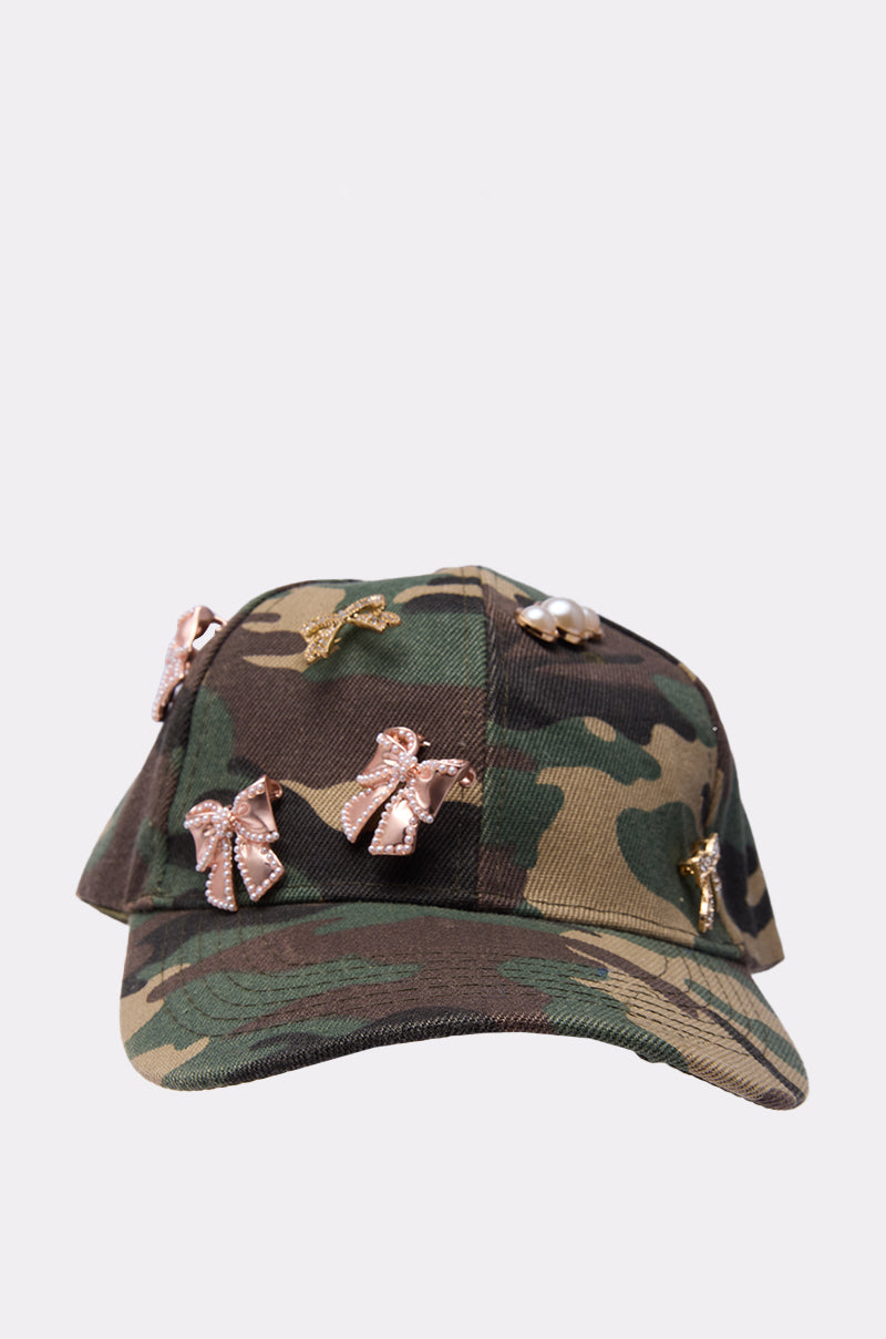 The TANYA CHARM HAT is a camouflage baseball cap with an adjustable velcro strap, featuring gold and pink jeweled brooches in bow, animal, and pearl designs, shown against a plain light background. 2
