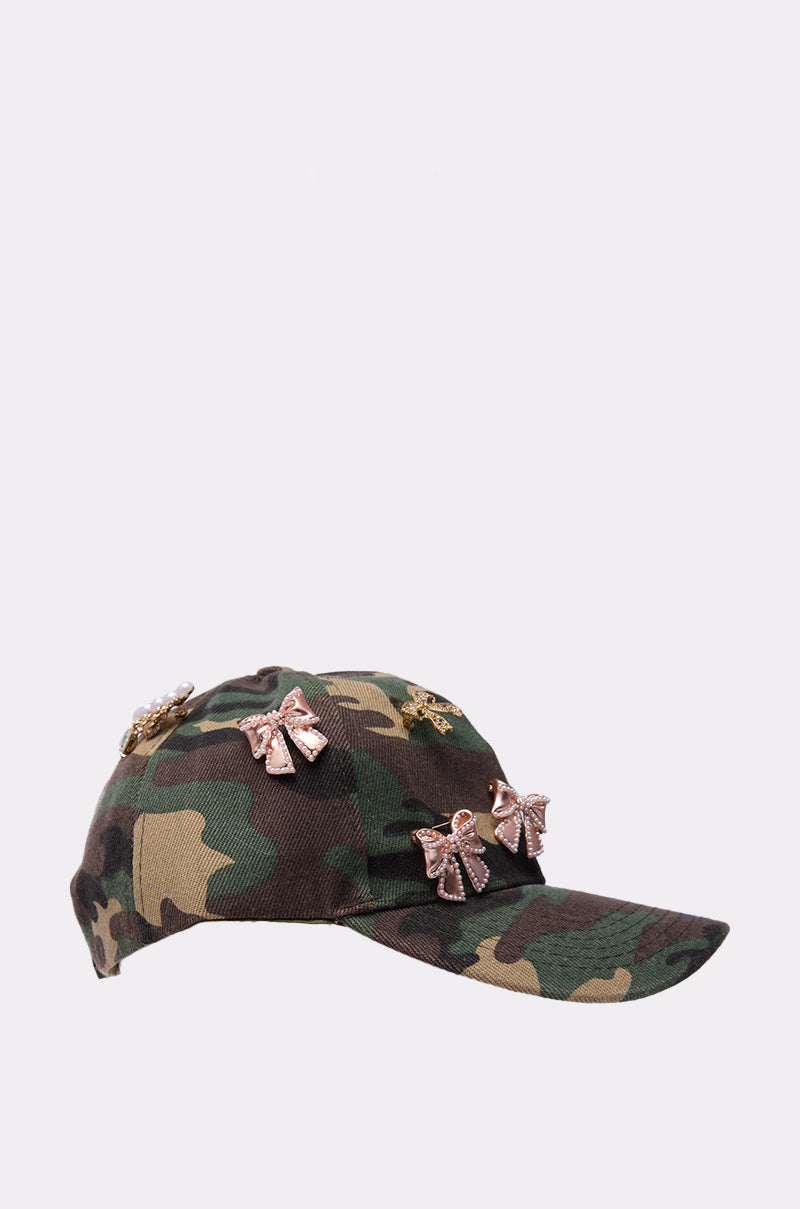 The TANYA CHARM HAT is a camouflage baseball cap adorned with multiple rose gold bow-shaped rhinestone accents, shown against a simple light backdrop. 3