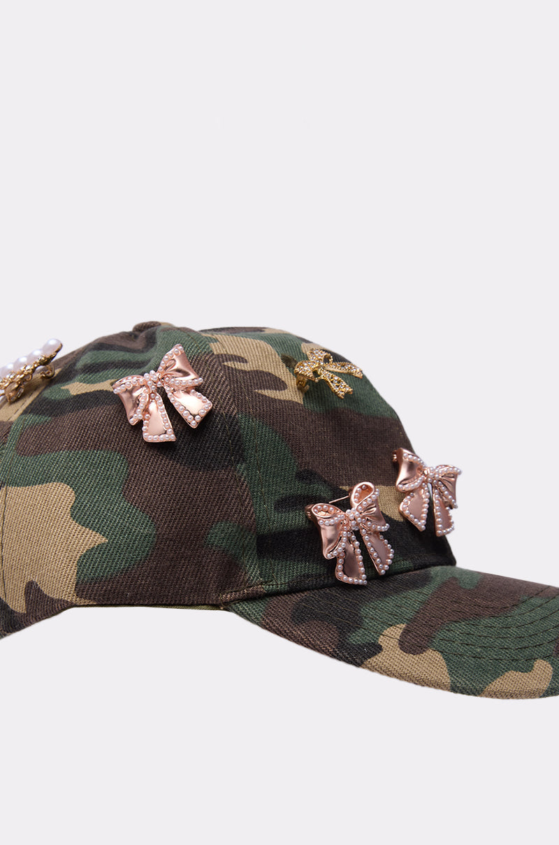 The TANYA CHARM HAT, a camouflage baseball cap embellished with metallic bow pins and sparkling rhinestones, stands out stylishly against a simple light background. 4