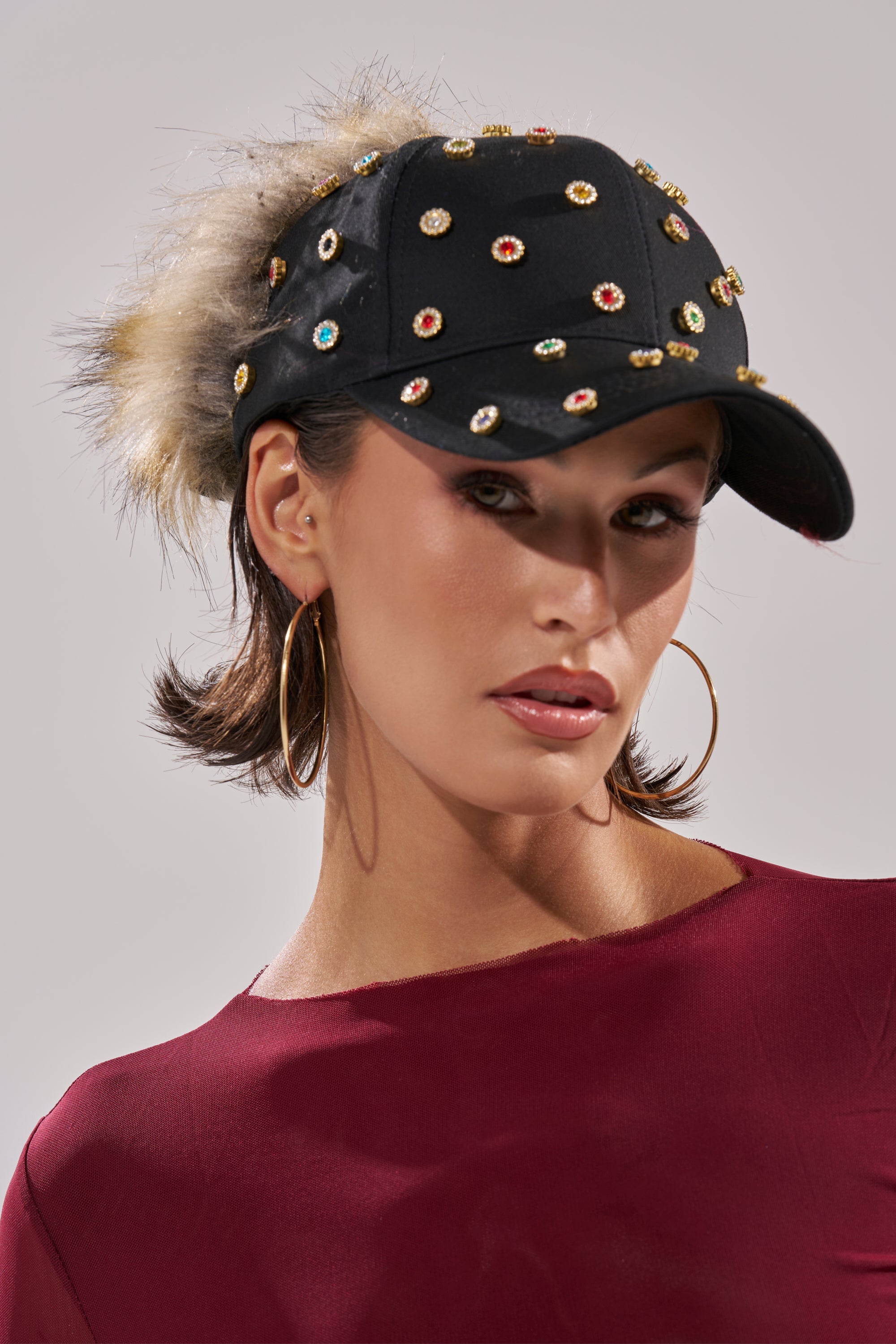 A woman wears the TJ FAUX FUR EMBELLISHED HAT with multicolor studs, paired with a maroon top and large hoop earrings, posing against a light gray background. 1