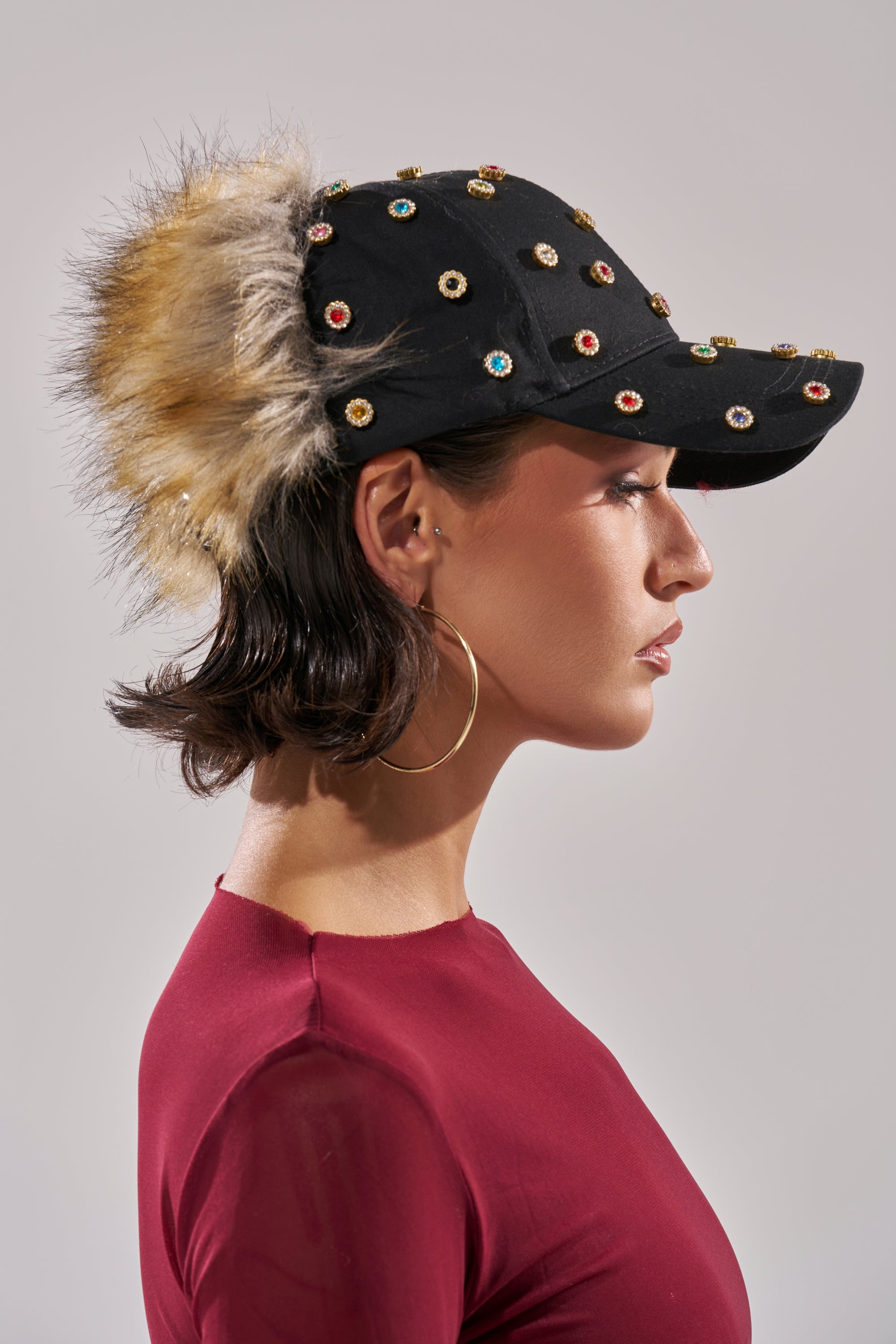 A person in a red top and large hoop earrings is shown in profile, wearing the TJ FAUX FUR EMBELLISHED HAT, featuring colorful jewels and faux fur trim against a plain, neutral background. 0