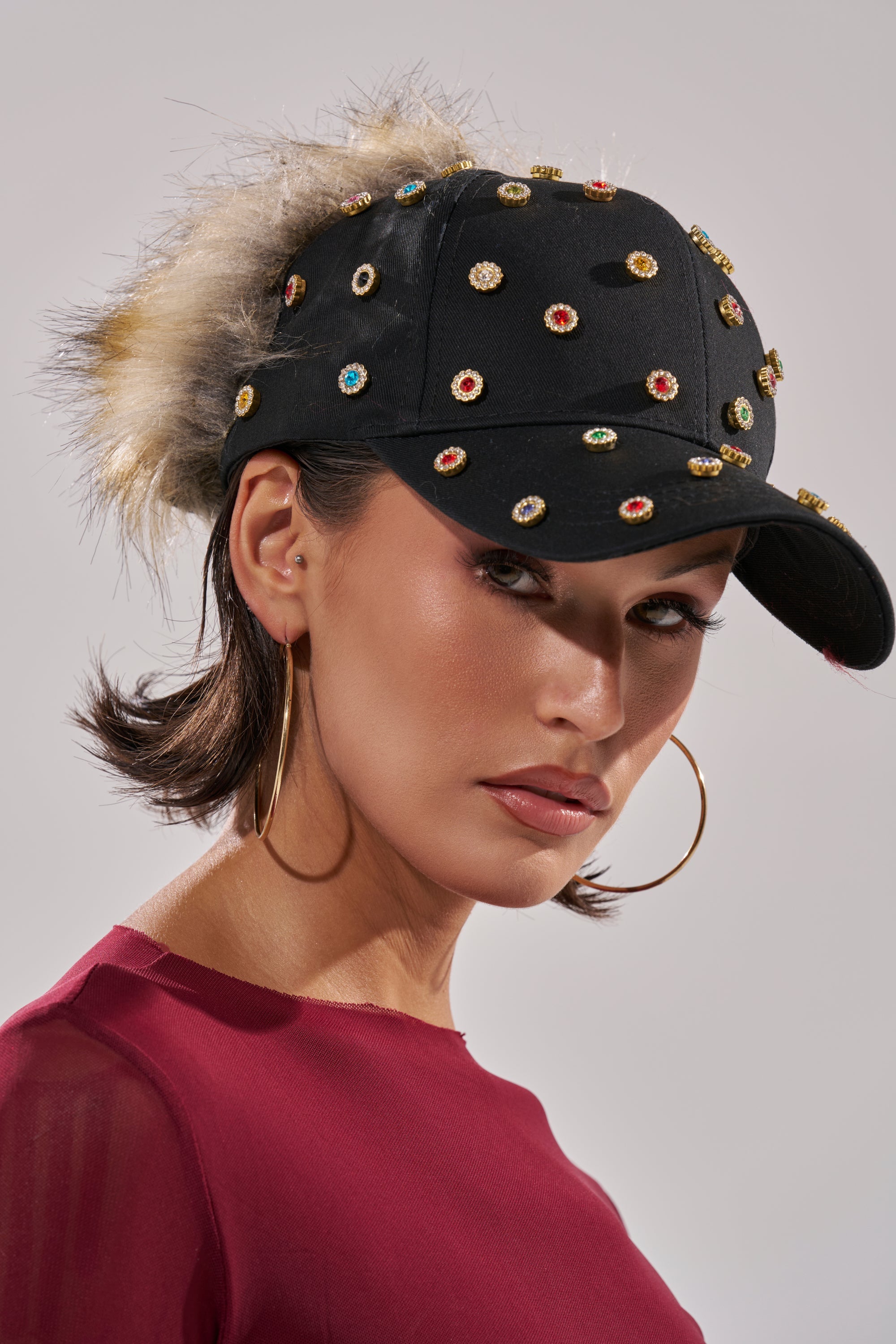 A woman confidently models the TJ FAUX FUR EMBELLISHED HAT, decorated with colorful gems and fur accents. She has short brown hair, large gold hoop earrings, and wears a sheer burgundy top. 2