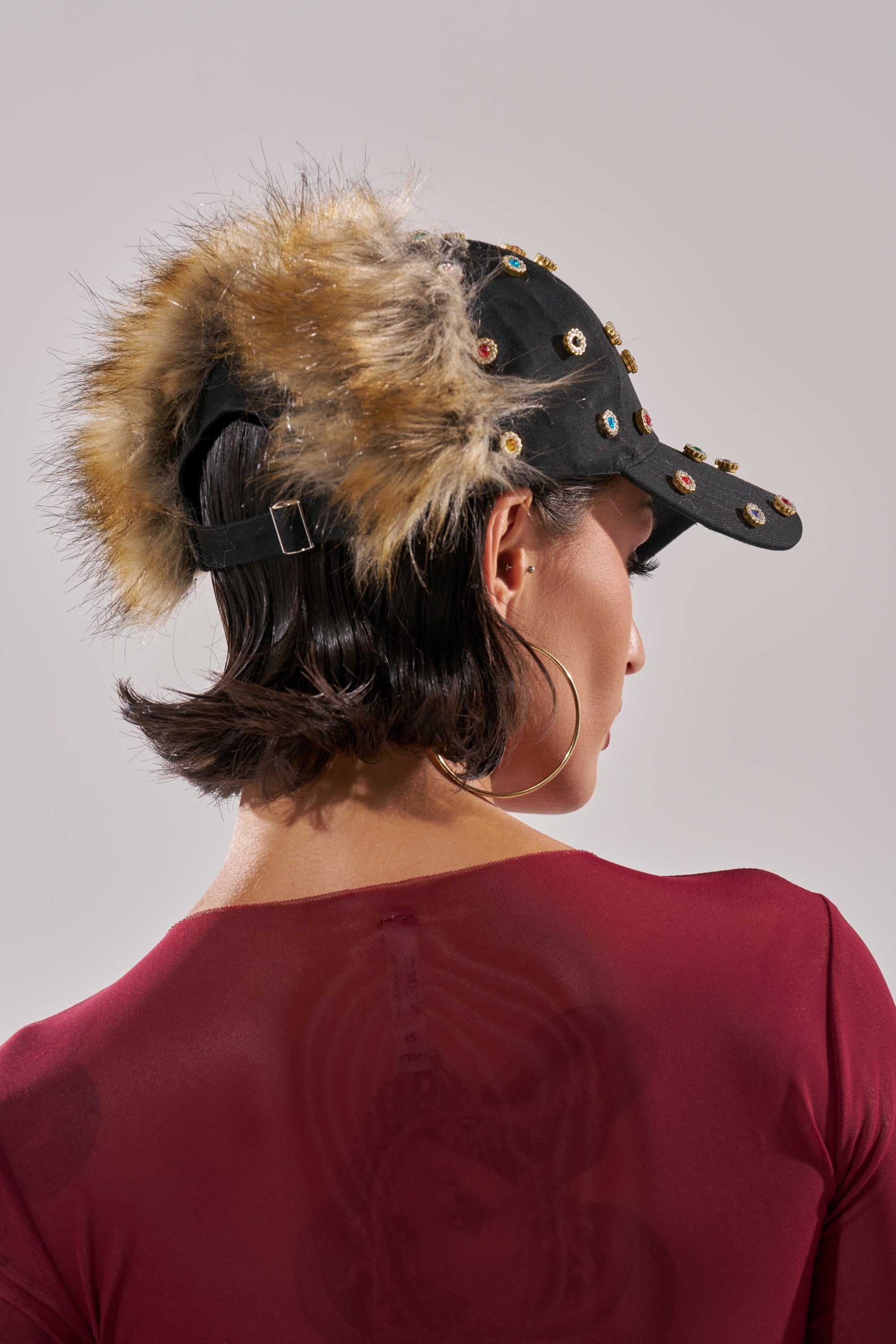 A person seen from behind wears the TJ FAUX FUR EMBELLISHED HAT, featuring fur trim and studs. They have short dark hair, large hoop earrings, and a sheer red top with a faint back image. 3