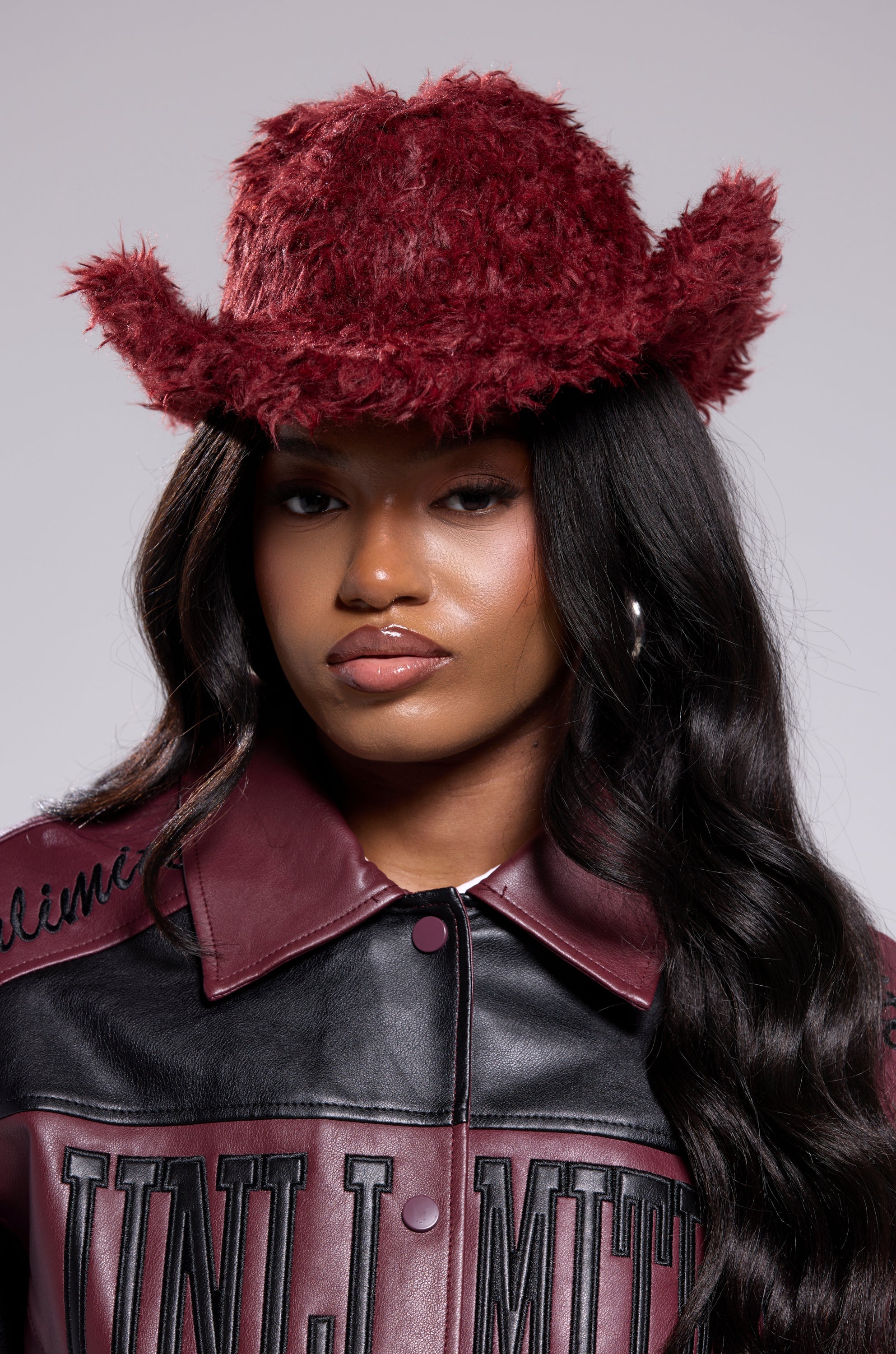 A woman with long wavy hair wears the FOR THE VIBES FAUX FUR COWBOY HAT in burgundy, paired with a matching burgundy and black "UNLMTD" jacket and hoop earrings, giving her look a bold Western flair. 2