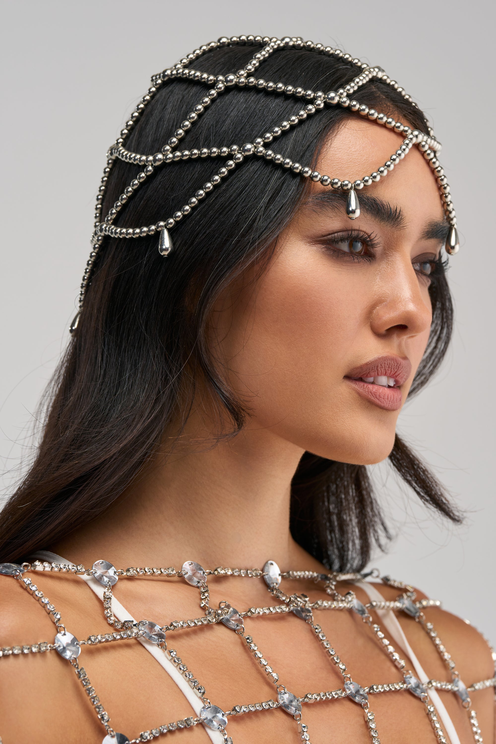 Woman with long dark hair wearing the WATERFALLS BEADED HEADPIECE, adorned with silver beads, and a matching grid-patterned top featuring crystal accents, looking to the side against a neutral background.