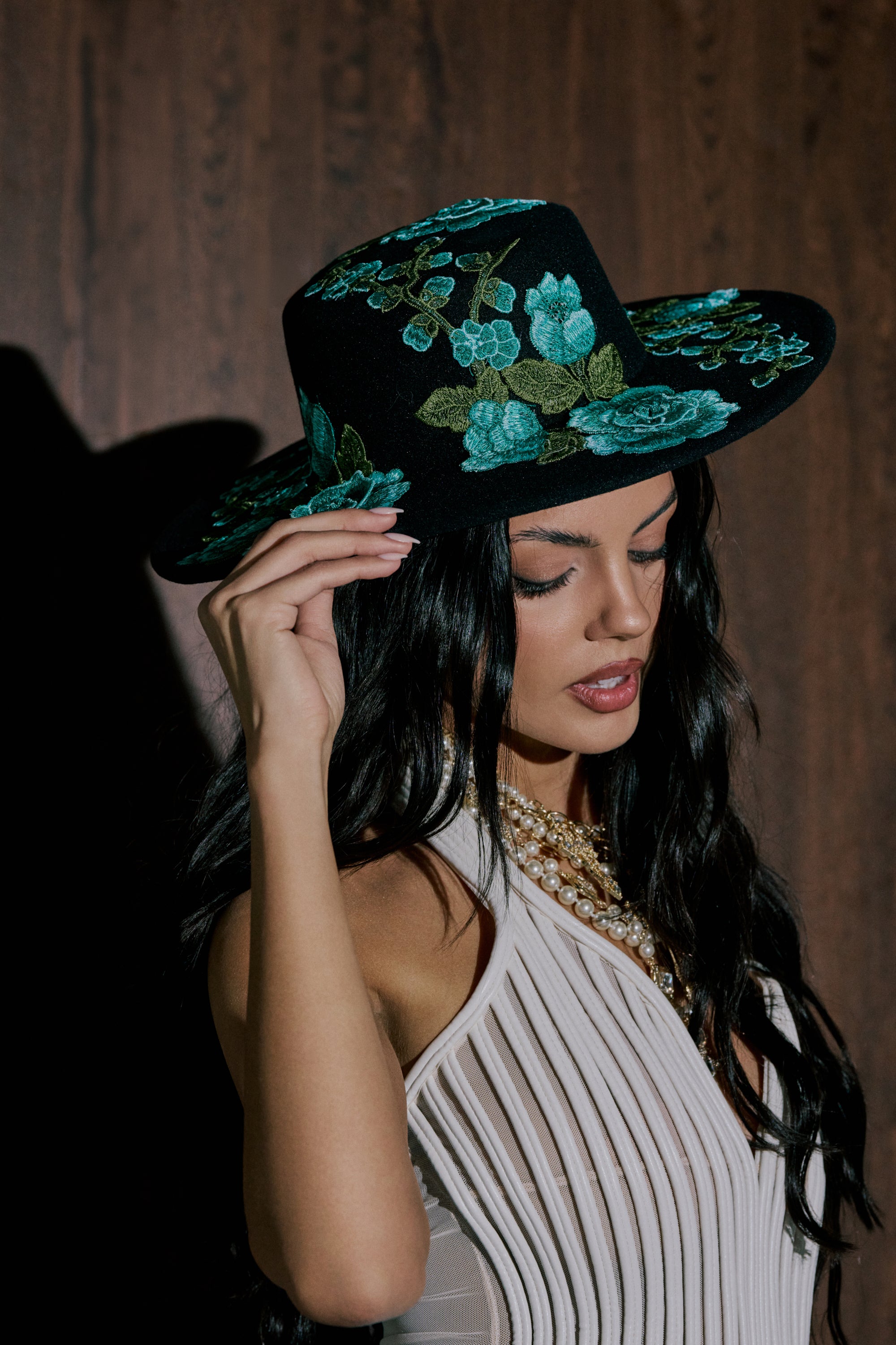 A woman with long dark hair and a pearl necklace wears a pleated white top and holds the IN THE FIELD APPLIQUE BRIM HAT, a statement wide-brim hat with blue and green embroidery, standing against a dark wooden background. 0