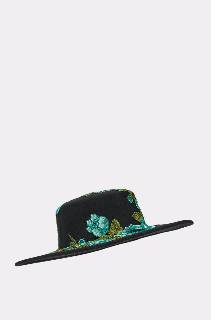 The IN THE FIELD APPLIQUE BRIM HAT is a black, wide-brimmed, flat-crowned statement piece featuring green and teal floral embroidery, shown against a plain light background. 3
