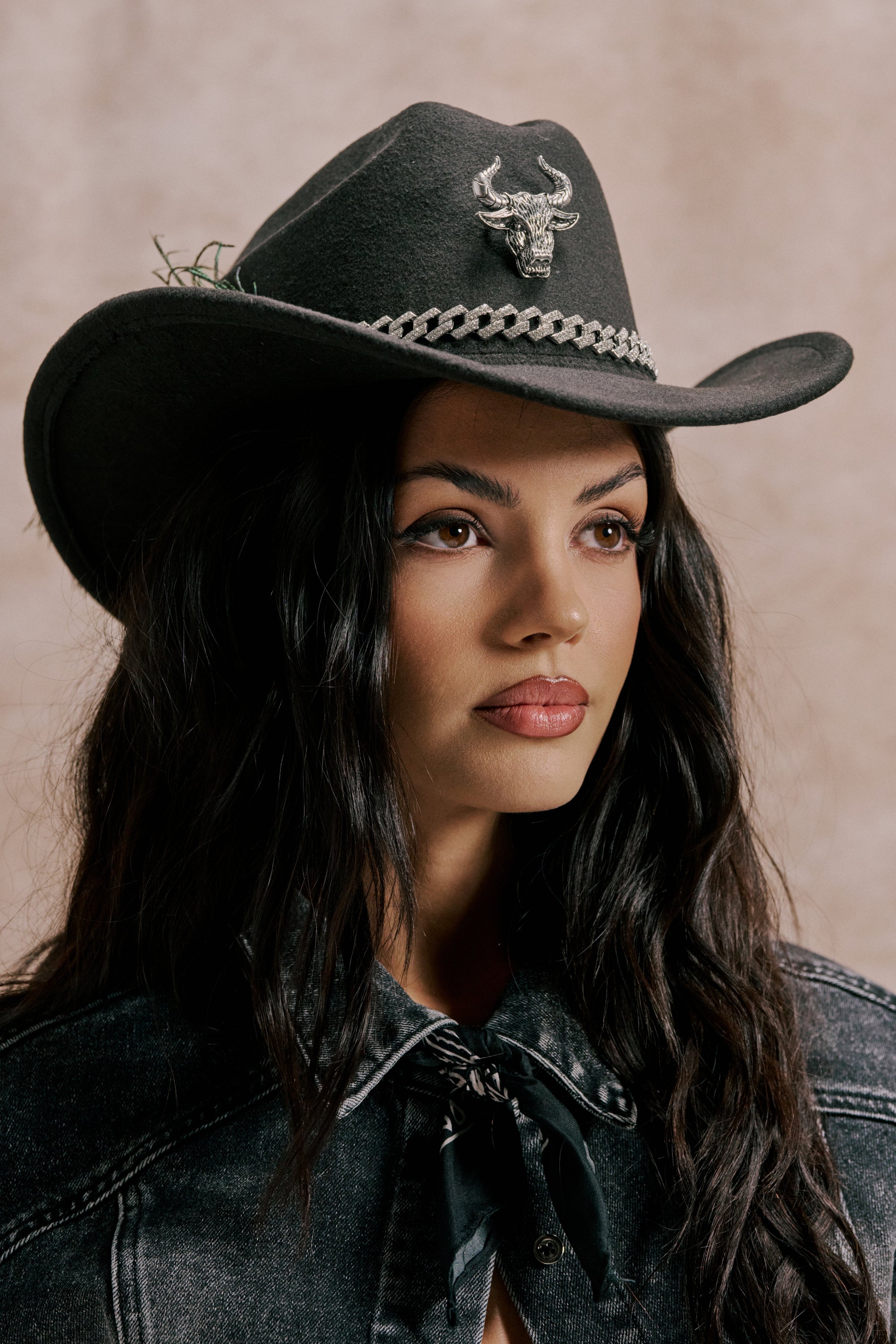 A woman with long dark hair wears the LETS RIDE COWBOY HAT, featuring a bull head pin and rhinestone chain detail, paired with a black denim jacket, as she looks slightly off-camera against a neutral background. 0