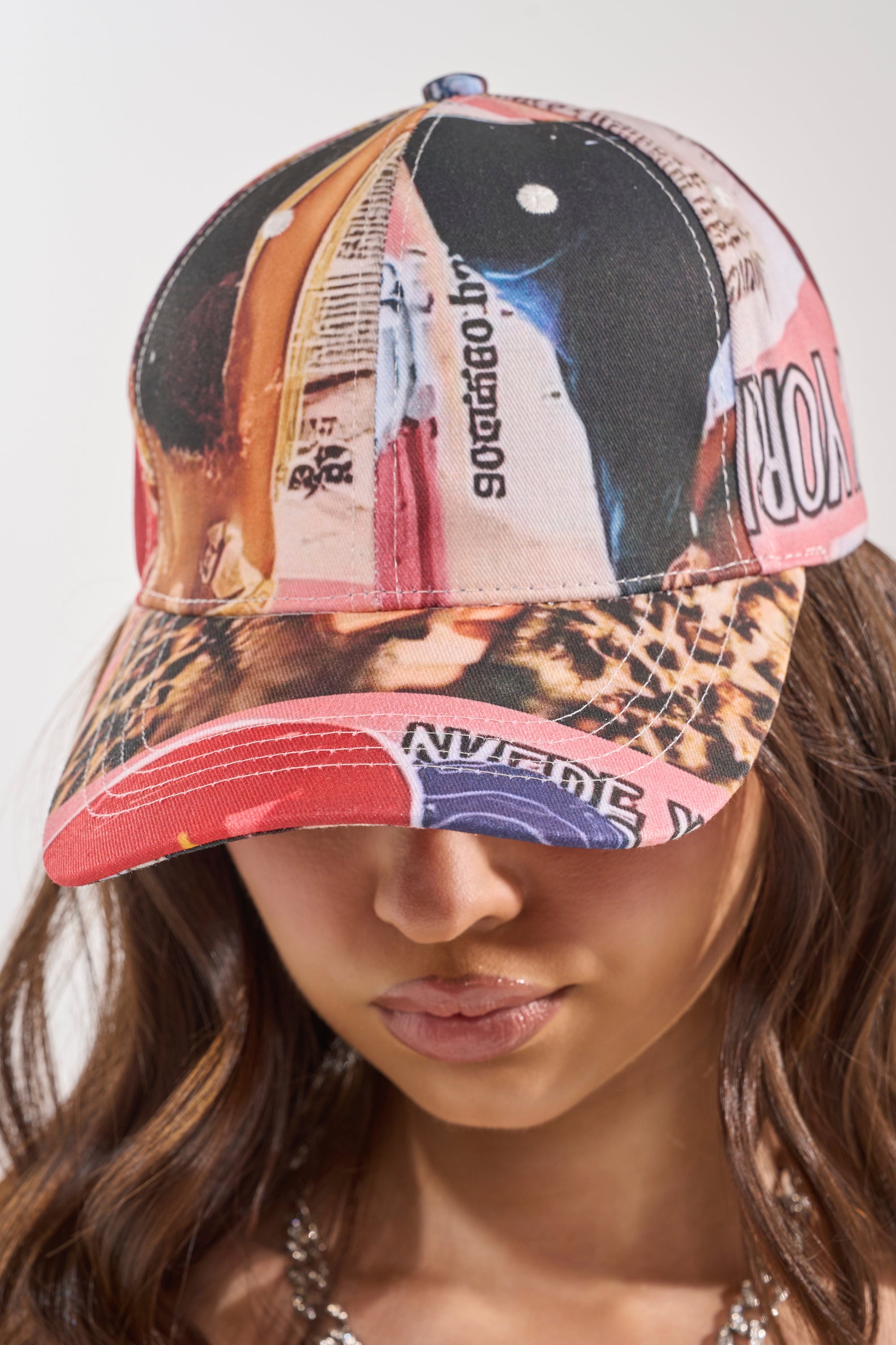 A person with long brown hair wears the IN THE ZINE PRINT HAT, a graphic baseball cap with an abstract collage design and text. The brim partly shades their eyes as they face forward against a neutral background. 2