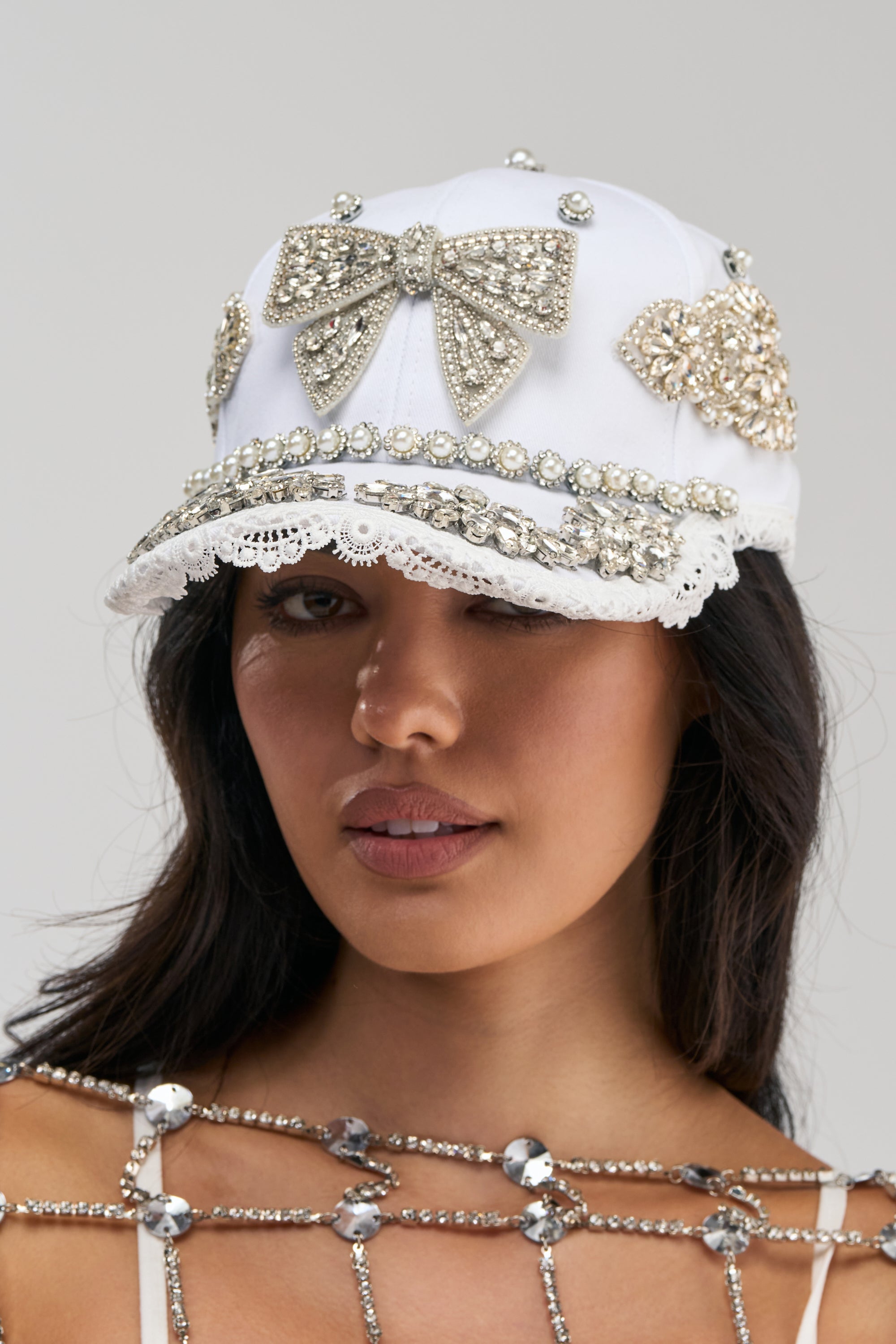A woman with long dark hair wears the IM OUTSIDE EMBELLISHED HAT, a white baseball cap adorned with large jeweled bows, pearls, rhinestones, and lace trim. Paired with her sparkling embellished top, this rhinestone and pearl hat is the perfect accessory for a bachelorette party.