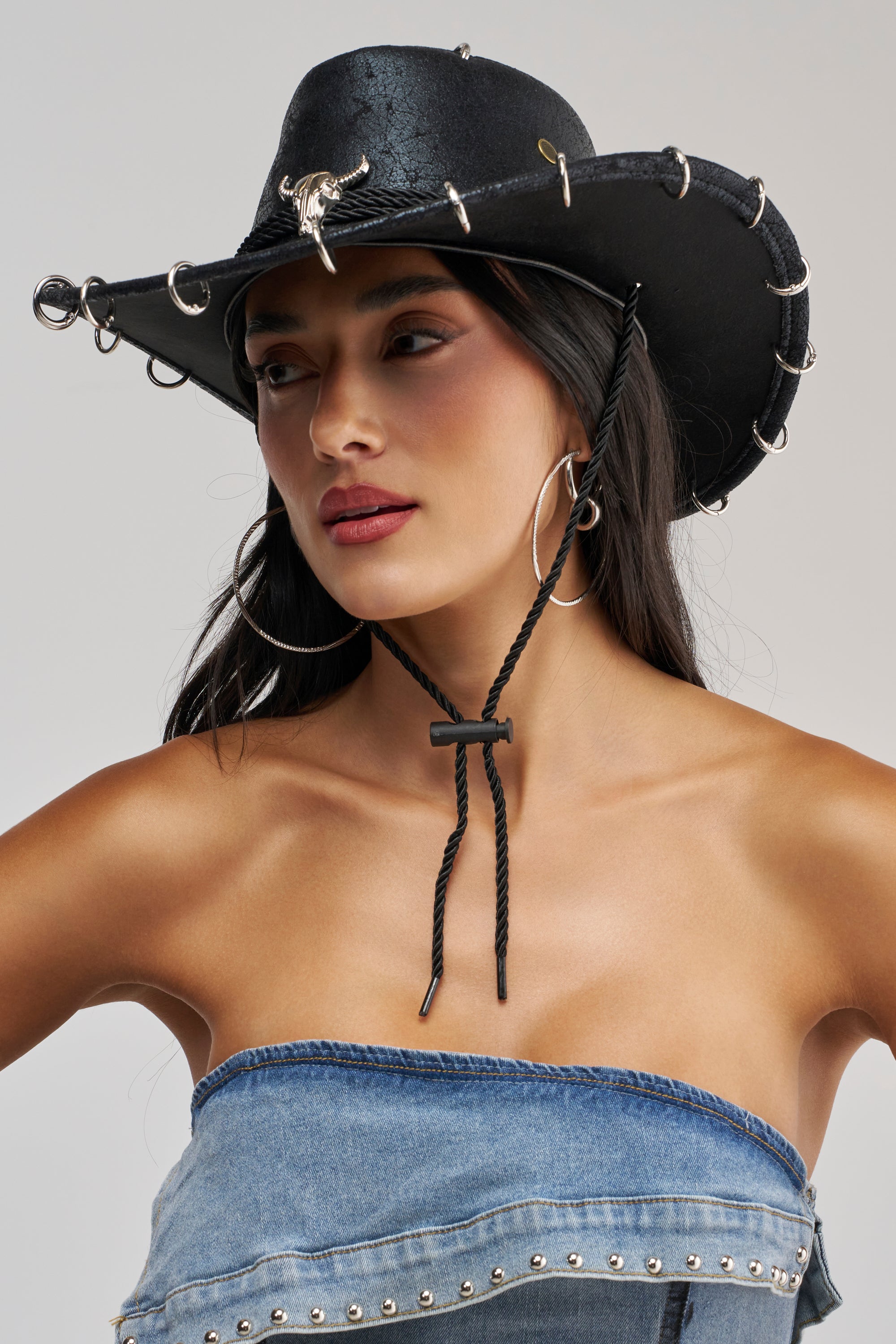 A woman models the TIME FOR THE RODEO COWBOY HAT—a black faux leather cowboy hat featuring silver rings and a bull emblem—paired with large hoop earrings and a strapless denim top, as she gazes slightly to the side against a neutral backdrop.