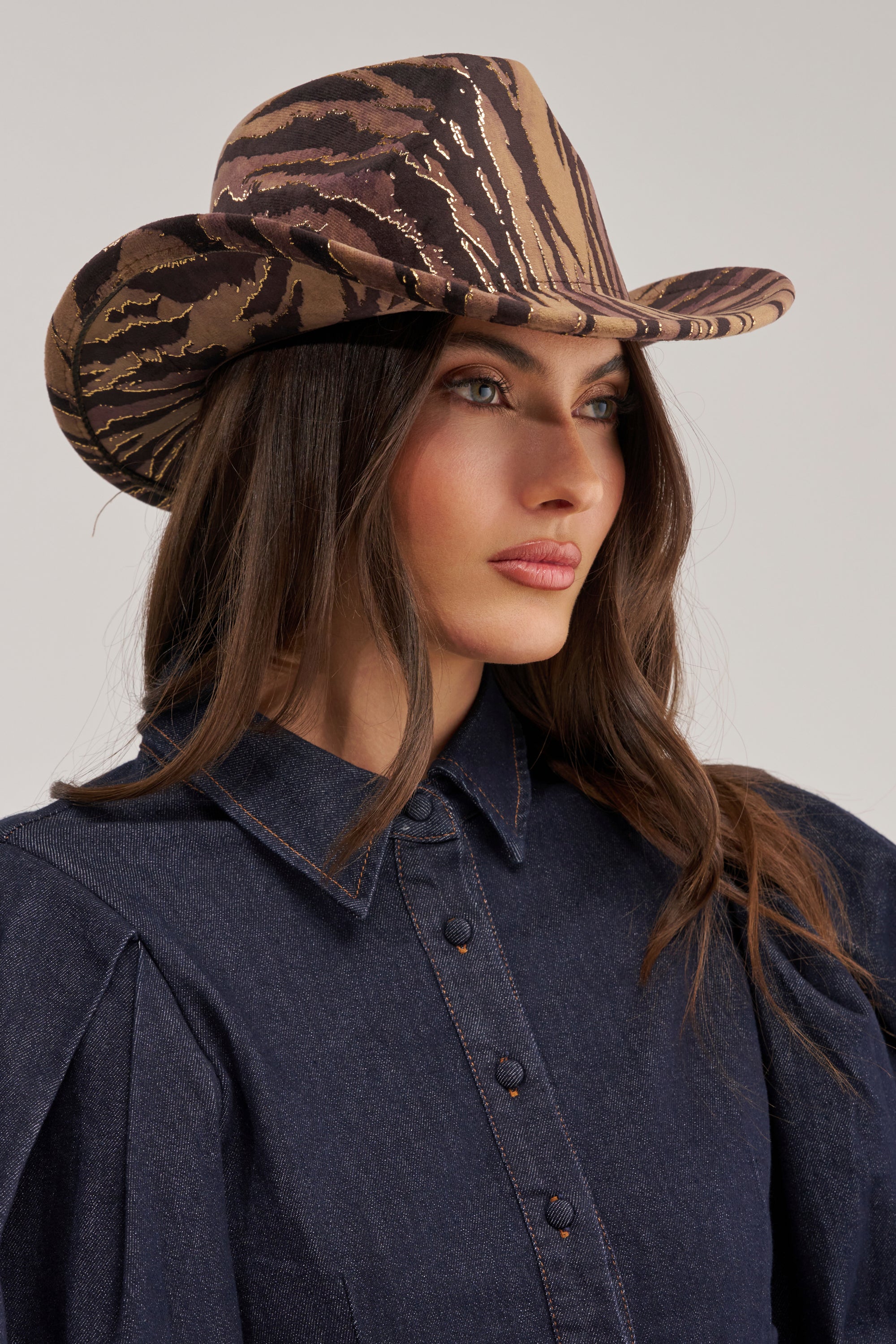 A woman with long brown hair wears the WILD ONE VELVET COWBOY HAT and a dark denim button-up shirt, looking to the side against a plain background.