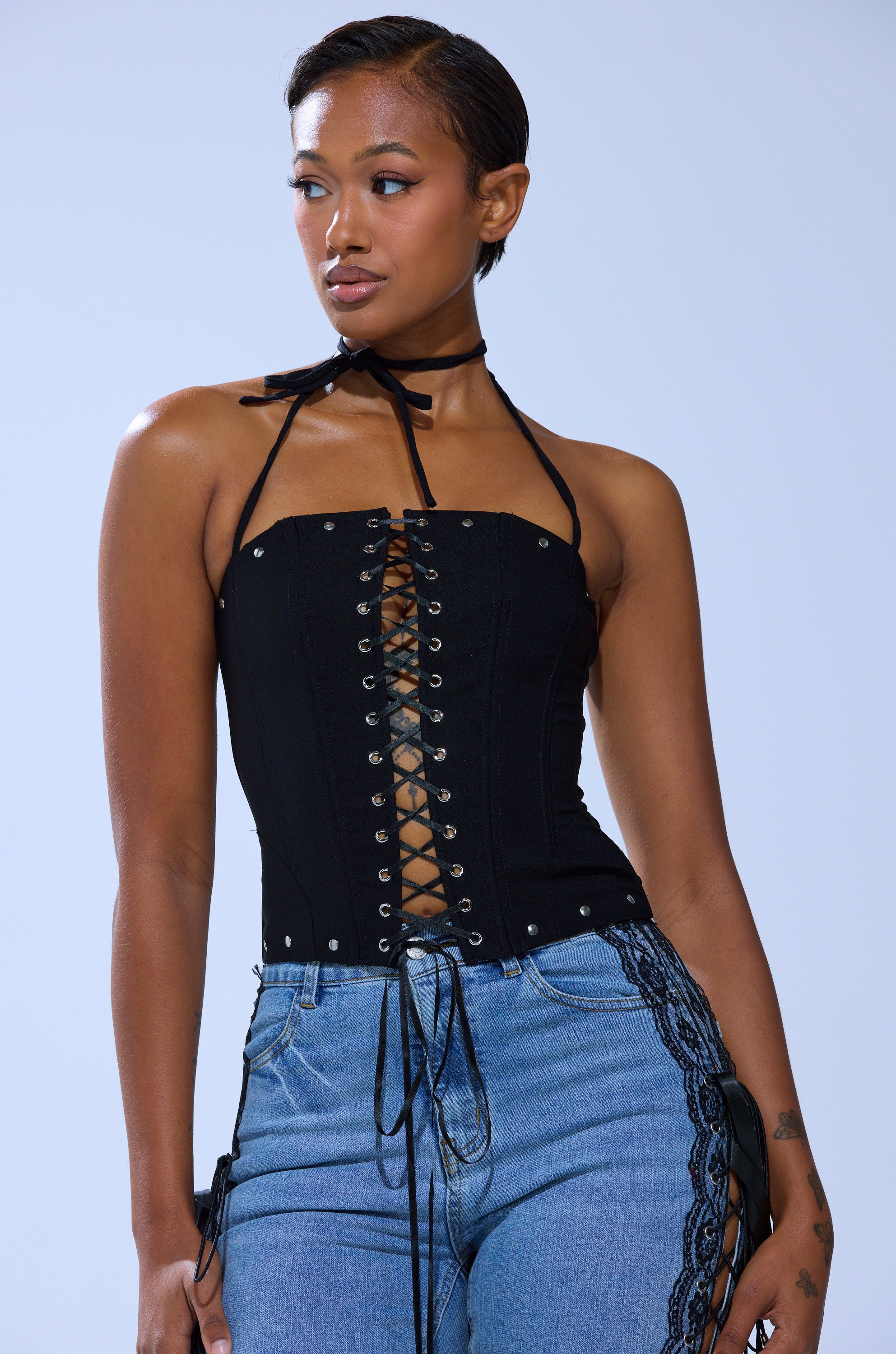 Wearing the MADISON LACE UP CORSET, a woman pairs the black lace-up halter top with stud details and blue jeans featuring black lace on the sides, posing against a plain light background.