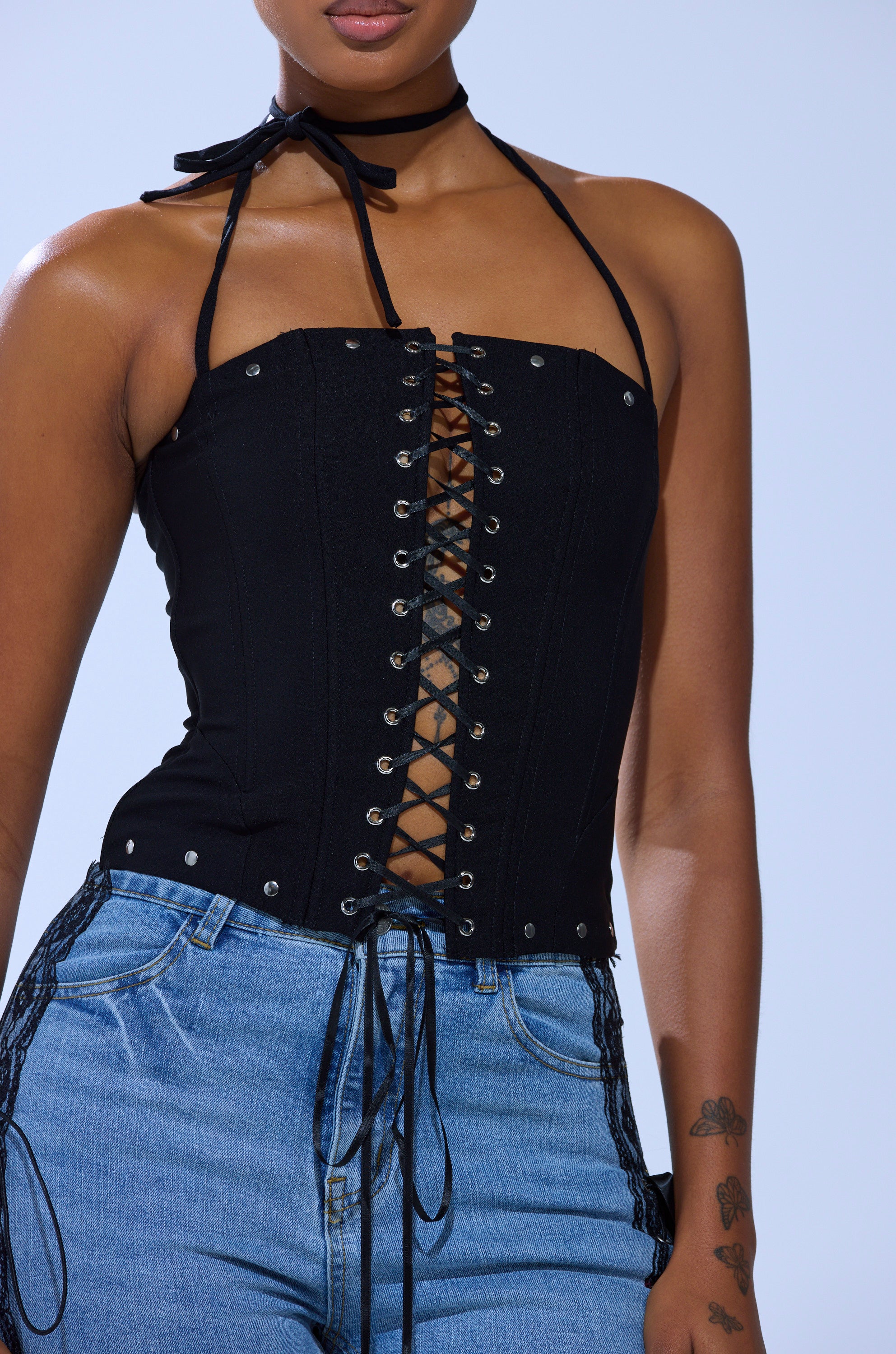 A person wears the MADISON LACE UP CORSET, a black lace-up halter top with silver grommets, styled with light blue jeans. The top ties at the neck, and their arm features small tattoos. They're set against a plain light blue background.