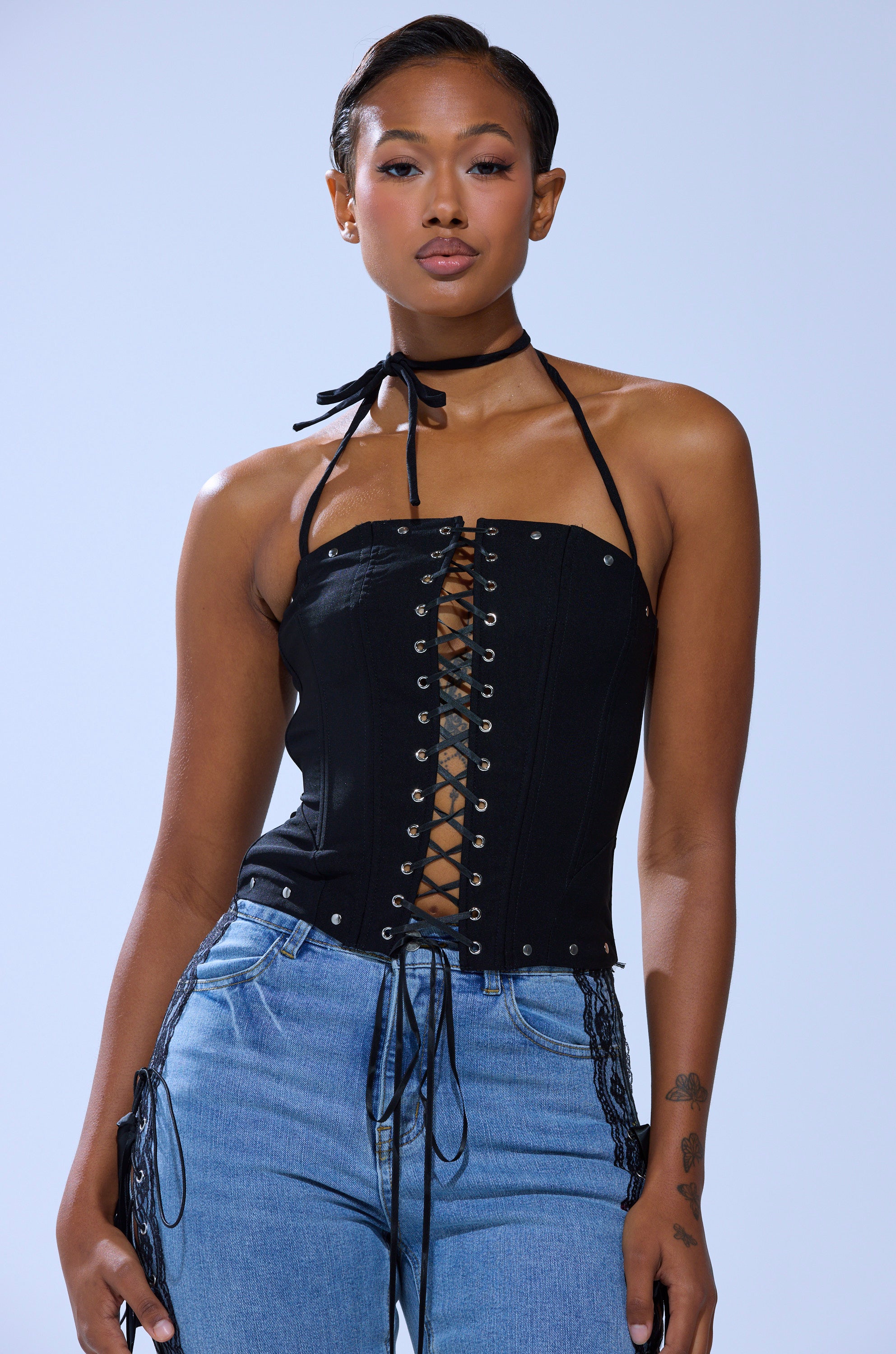 A person stands facing forward, wearing the MADISON LACE UP CORSET—a black corset top with silver stud details—paired with blue jeans. Their hair is neatly styled and they look confident.