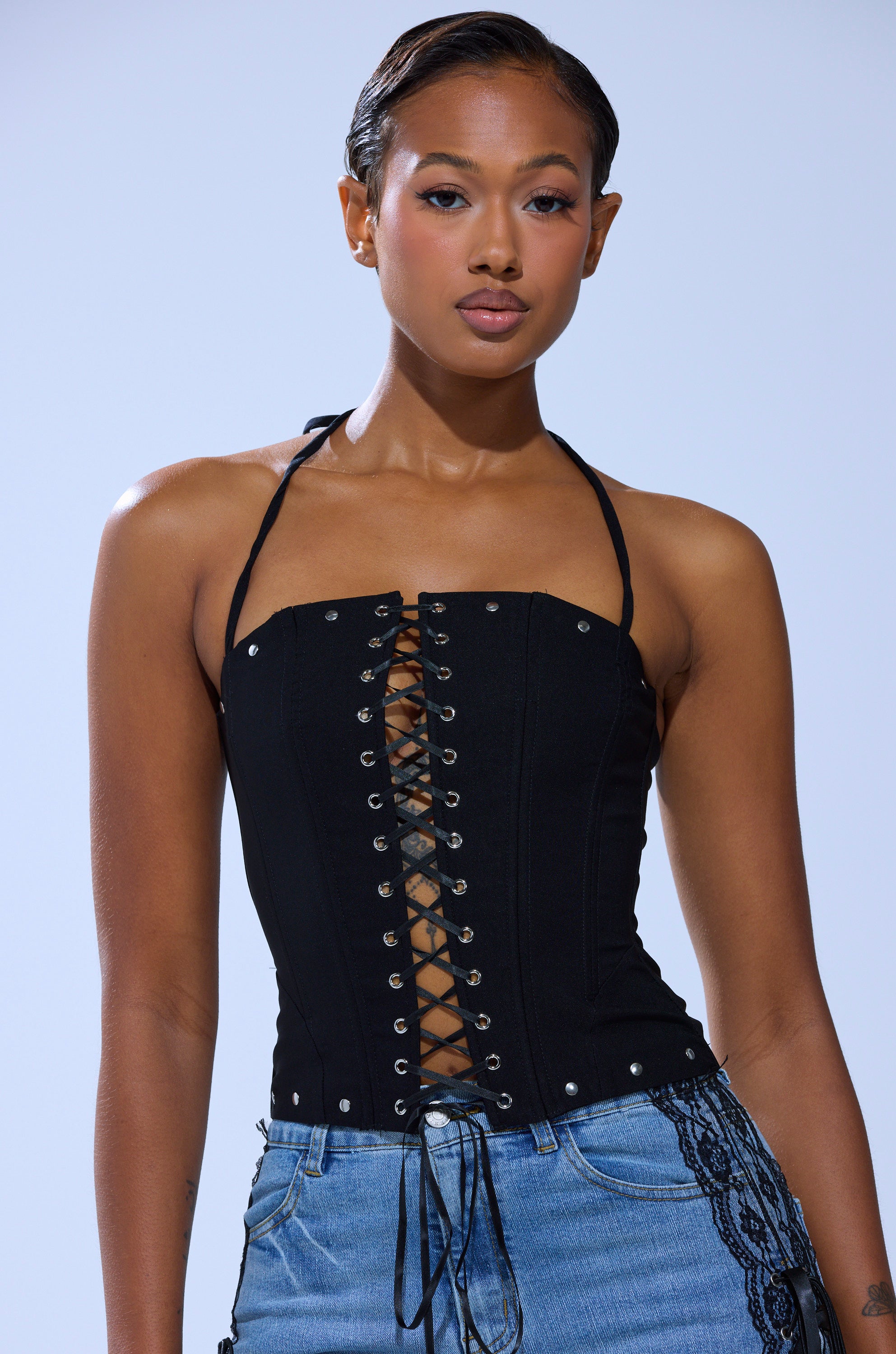 A woman wearing the MADISON LACE UP CORSET with silver eyelets and blue jeans stands against a plain light background, confidently facing the camera. The edgy lace-up detailing elevates her timeless style.