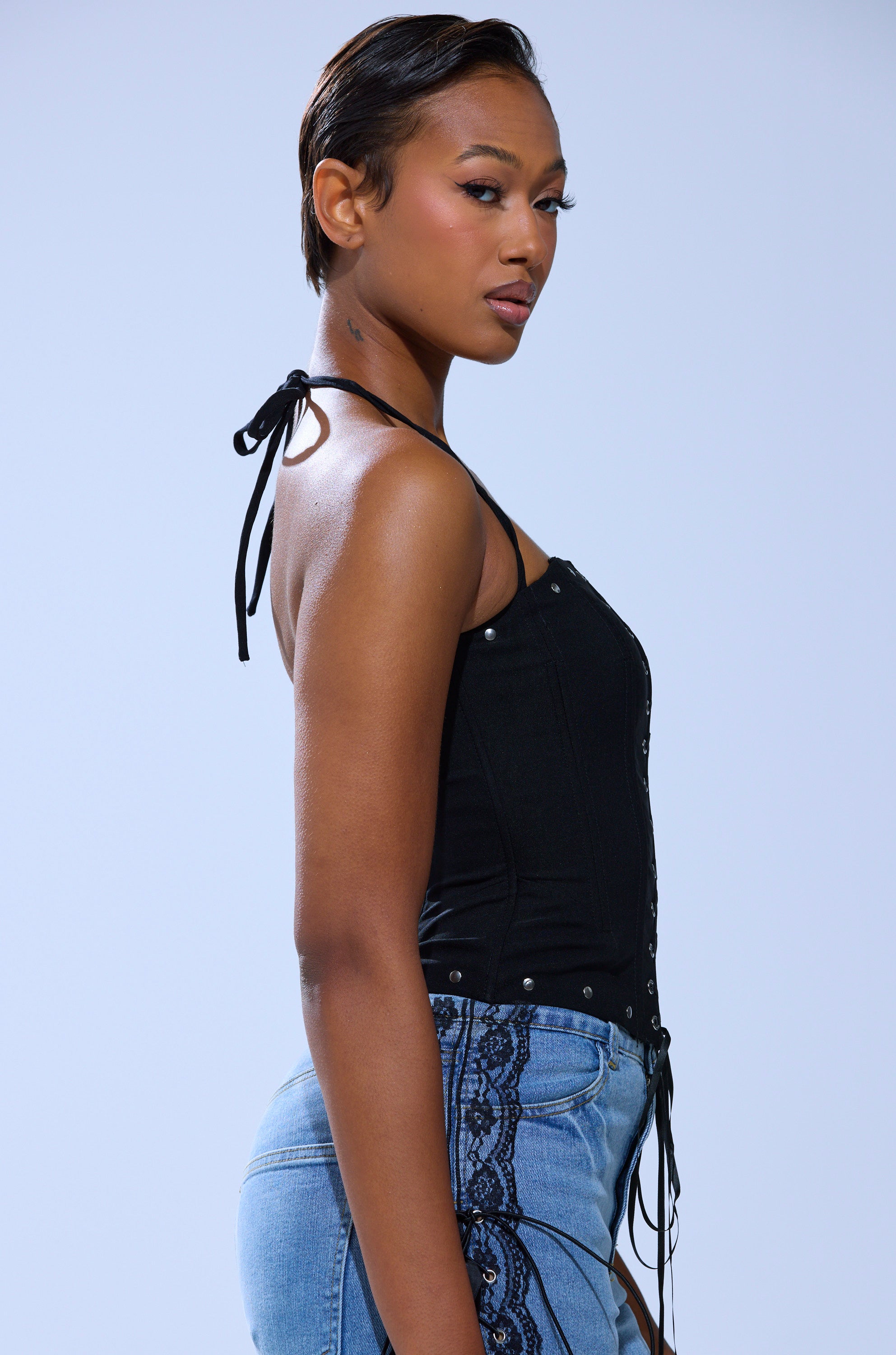 A woman with short hair poses in profile, wearing the MADISON LACE UP CORSET halter top and blue jeans with black lace-up corset sides, set against a plain light background.
