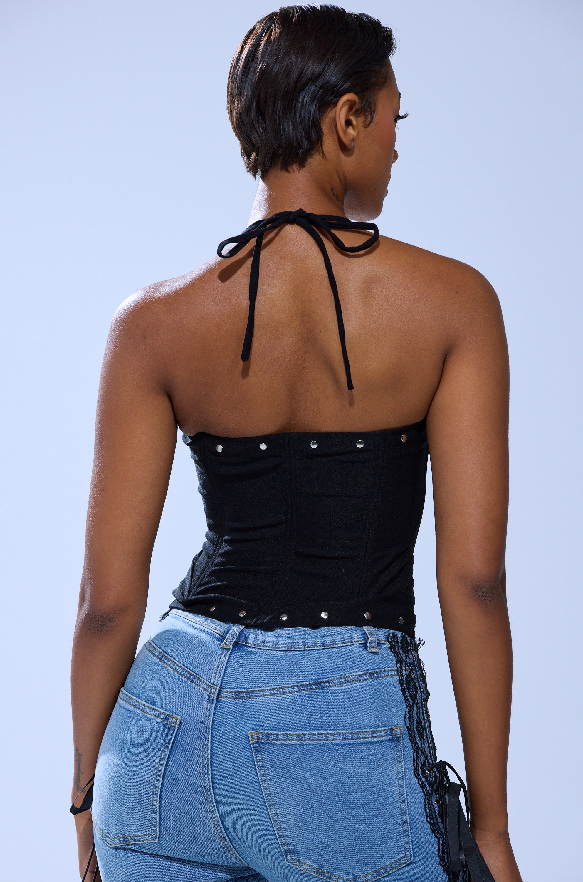 A woman with short hair is shown from the back, wearing the MADISON LACE UP CORSET in black with silver stud details and light blue jeans, standing against a plain light-colored background.