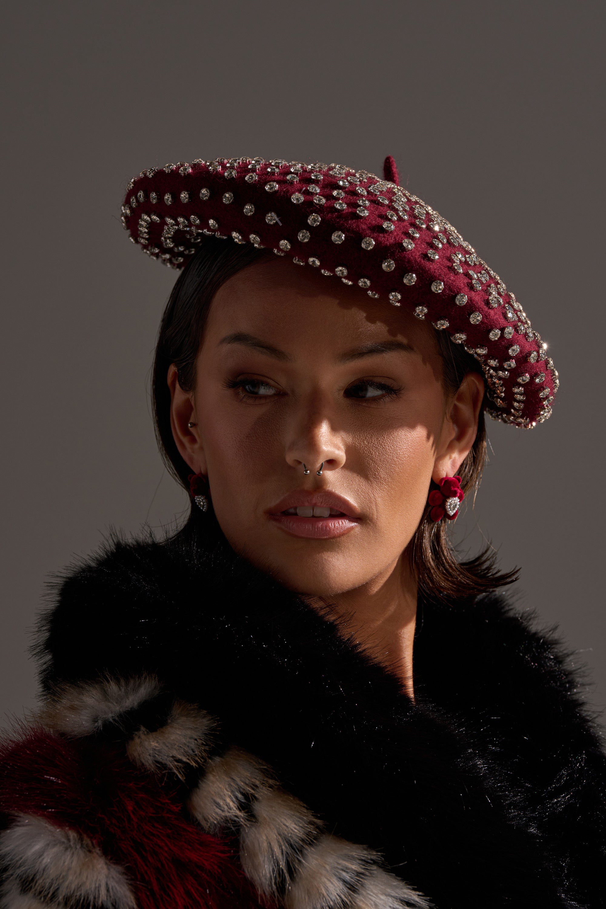 A person wears THE REAL RELATE EMBELLISHED BERET with rhinestone details, matching earrings, and a black fur coat with patterned accents, gazing slightly to the side against a neutral background. 1