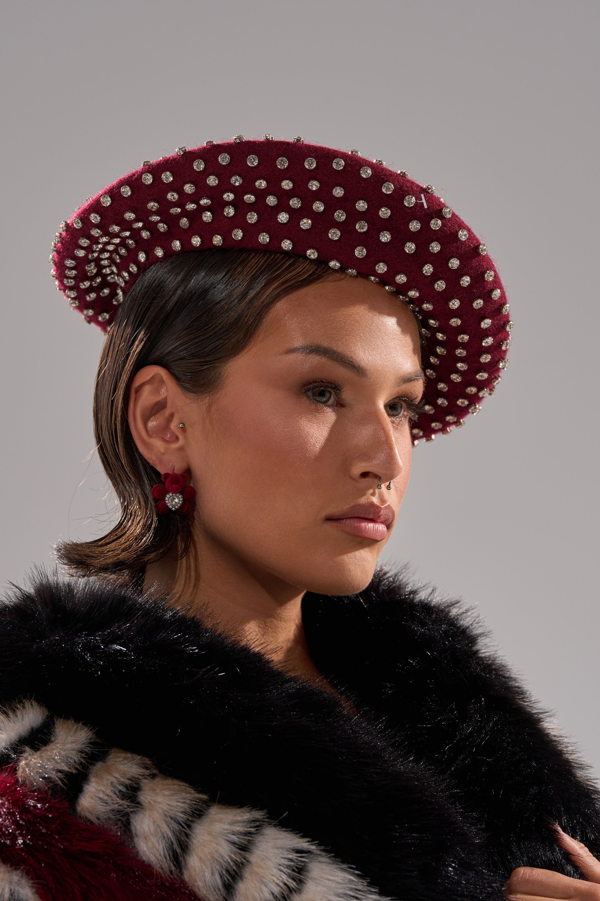 A person wearing THE REAL RELATE EMBELLISHED BERET, decorated with pearls, pairs it with red earrings and a black-and-white faux fur garment while gazing to the side against a neutral background. 2