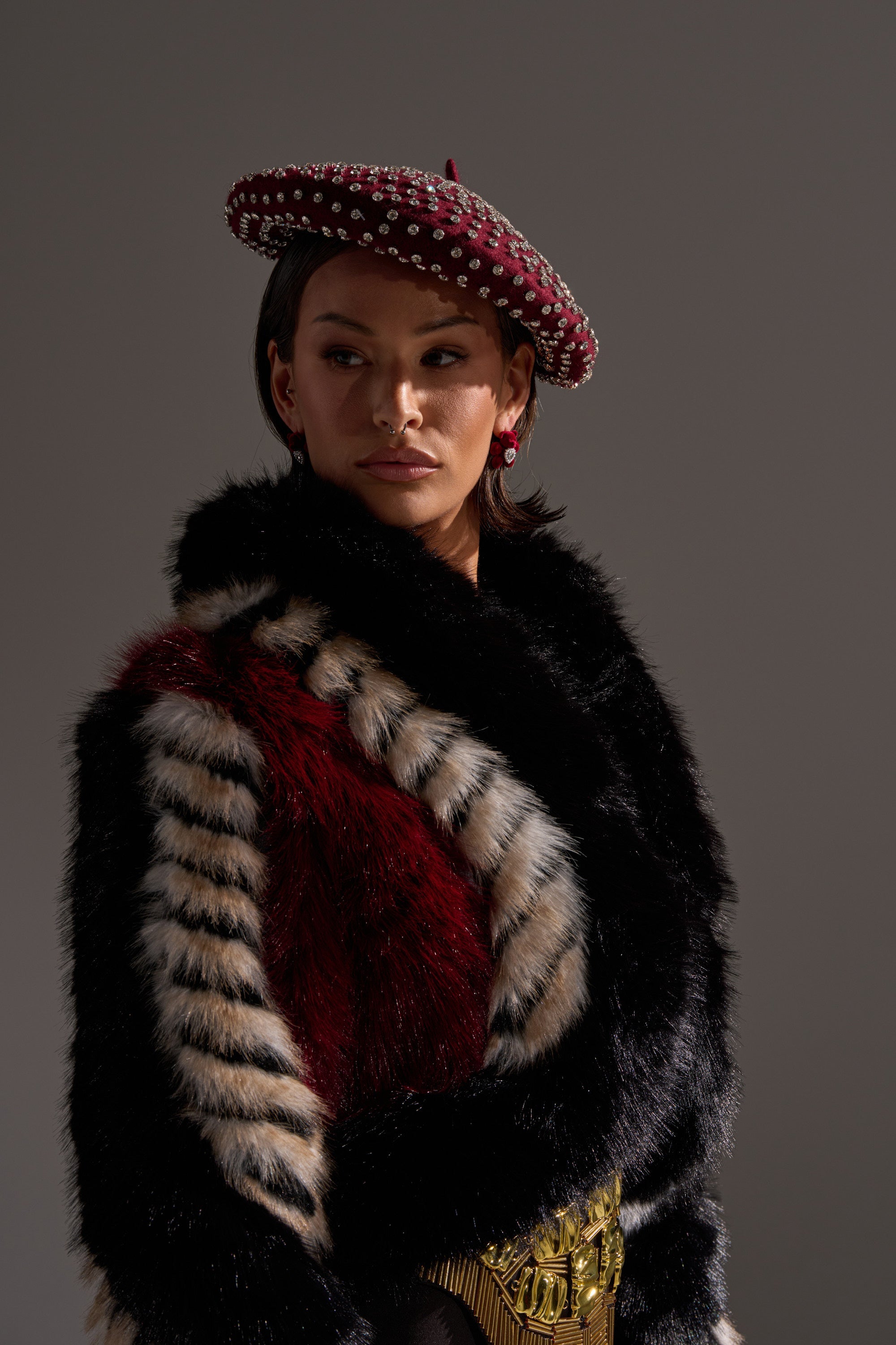 A person wearing a black, white, and red patterned fur coat and THE REAL RELATE EMBELLISHED BERET—a maroon wool beret with pearls—stands against a gray background, looking to the side. 6