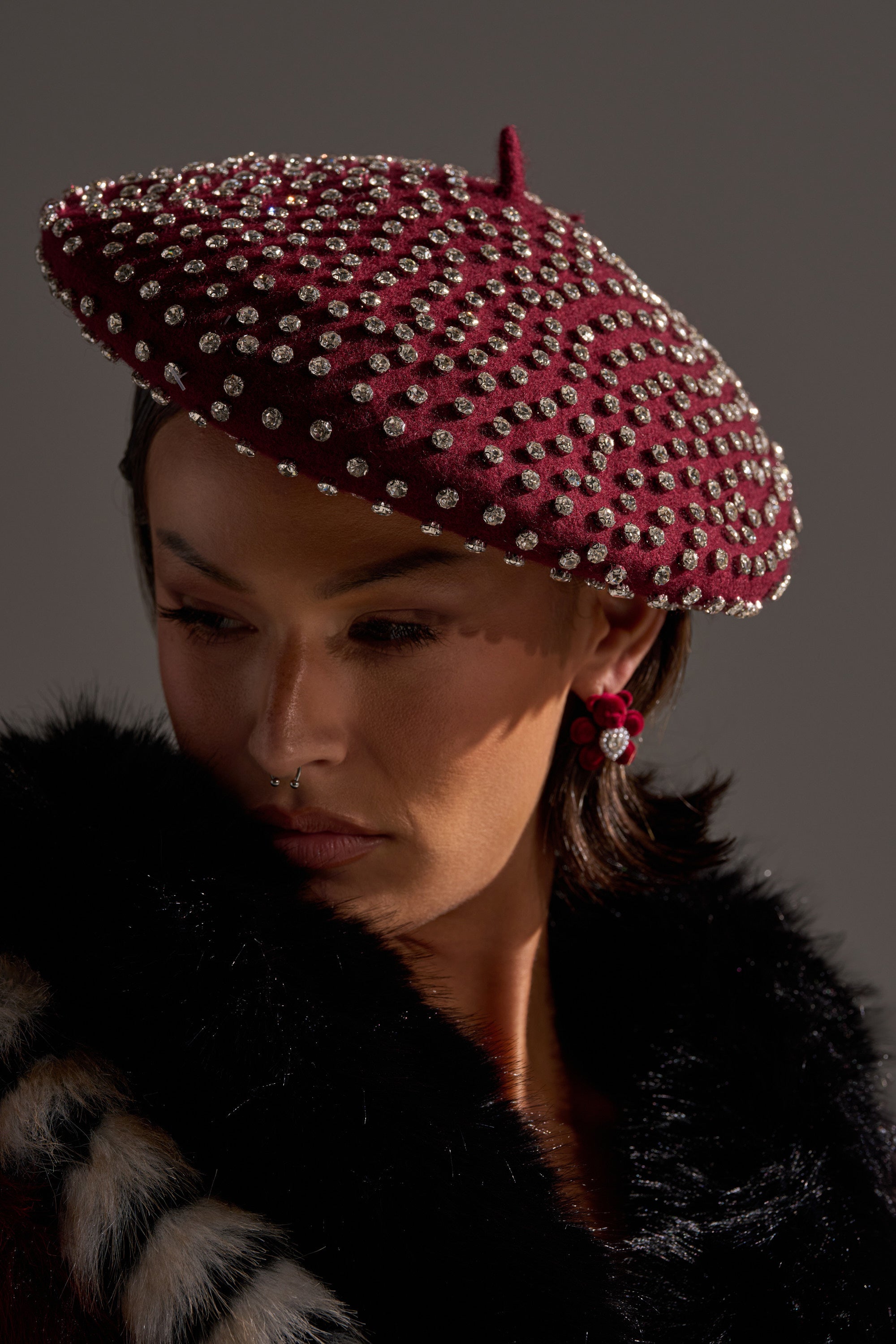 Wearing THE REAL RELATE EMBELLISHED BERET, a person in a dark fur coat with bold earrings and a septum piercing looks down, dramatic lighting casting shadows across their face. 7