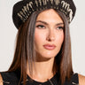 A woman with straight, brown hair wears the SAFETY FIRST BERET from AKIRA Label, accented with silver safety pins. She is also dressed in a coordinating black top featuring similar safety pin embellishments. She gazes forward with a neutral expression against a plain background.